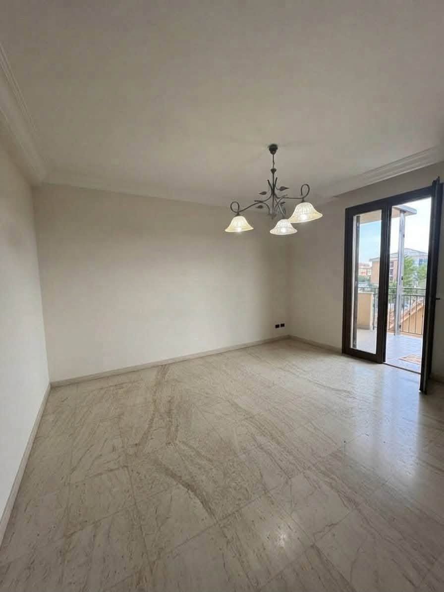 Sala da pranzo moderna e vuota con pavimento in legno chiaro, lampadario e porte scorrevoli in vetro che conducono a un balcone.