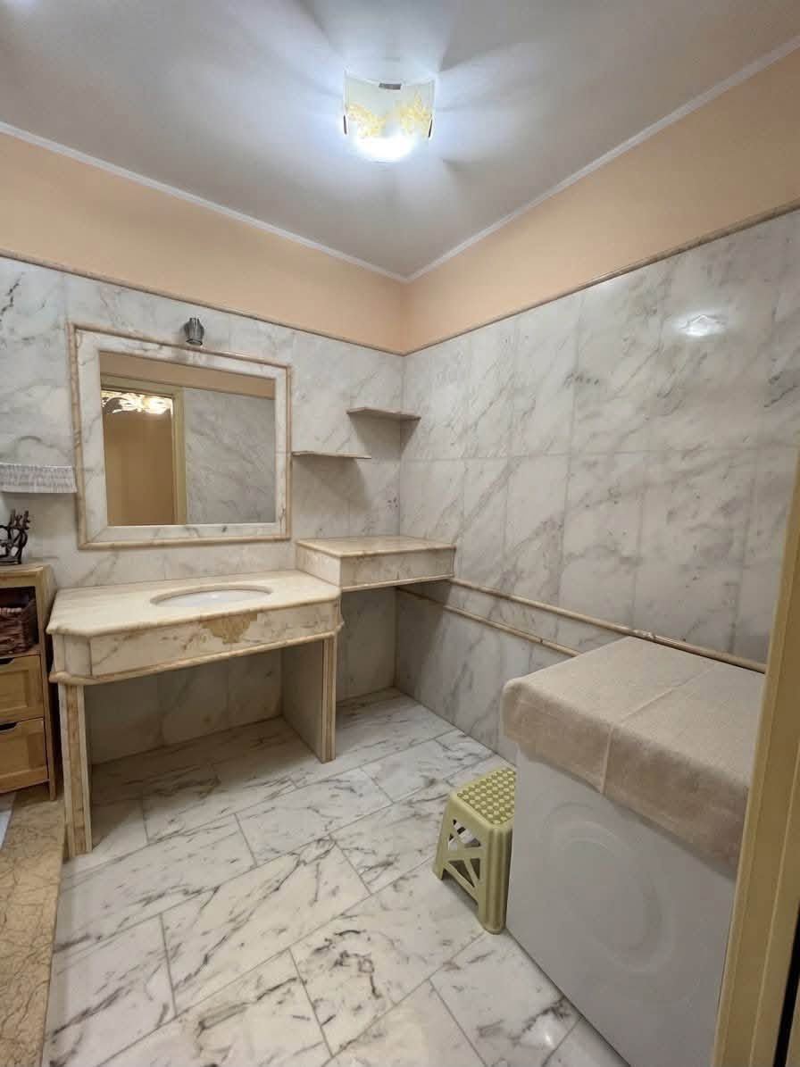 Piccolo bagno piastrellato in marmo con lavabo, specchio, mensole e WC sotto una lampada a soffitto.
