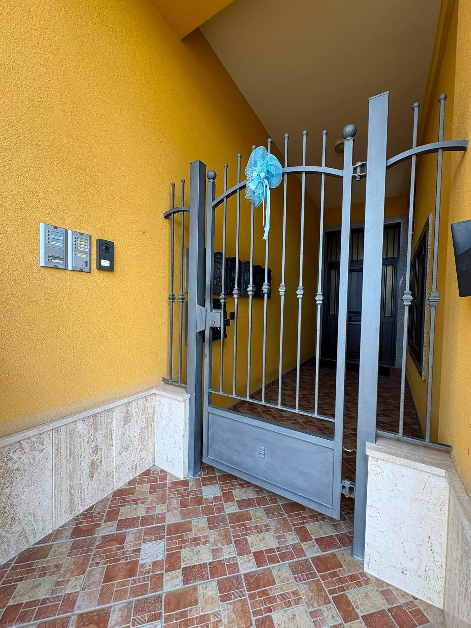 Ingresso con cancello in metallo decorato con nastro blu, pareti gialle e pavimento piastrellato in uno stretto passaggio.