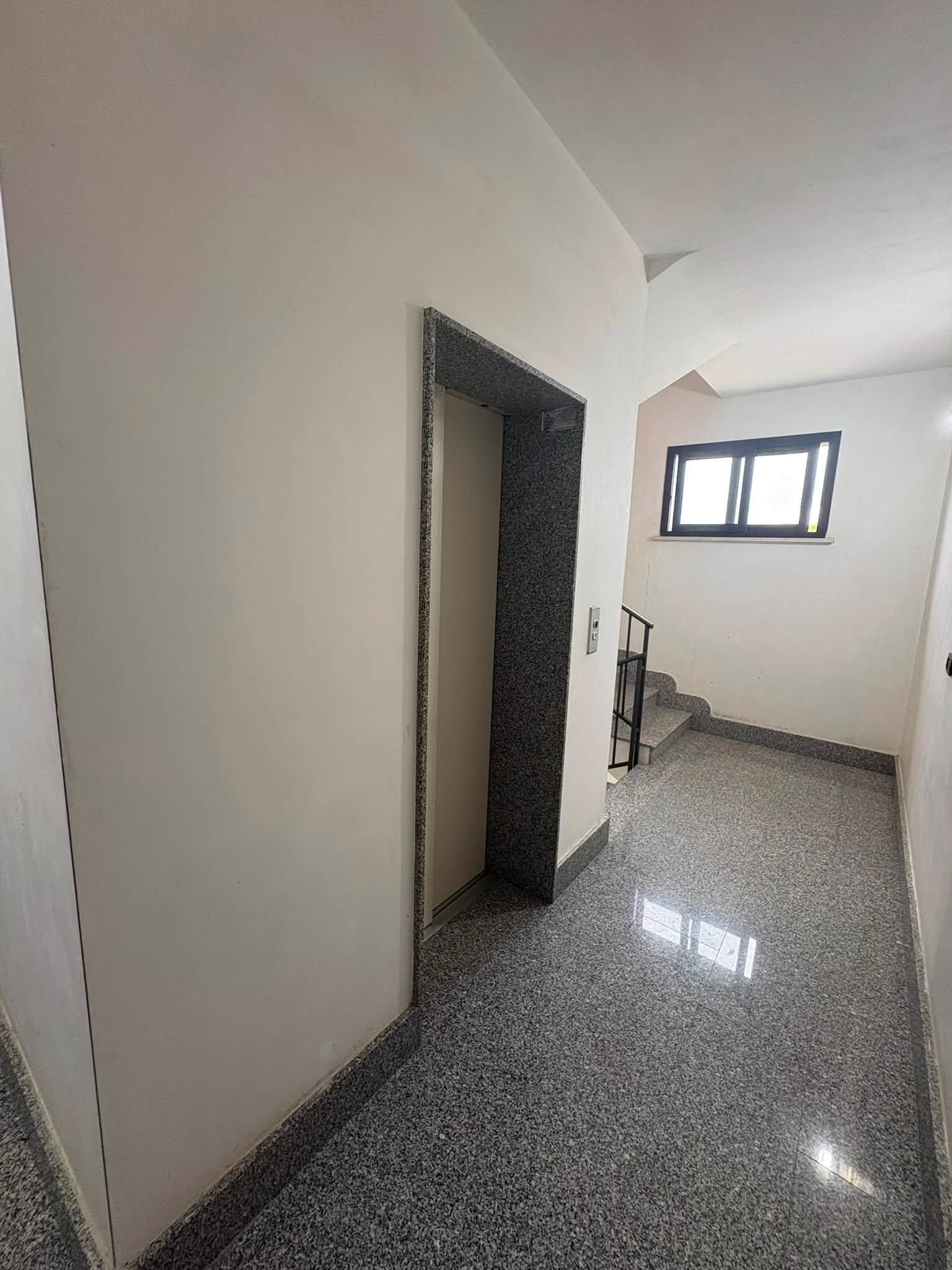 Corridoio vuoto con pareti bianche, pavimento piastrellato a chiazze, porta aperta e piccola finestra in fondo.