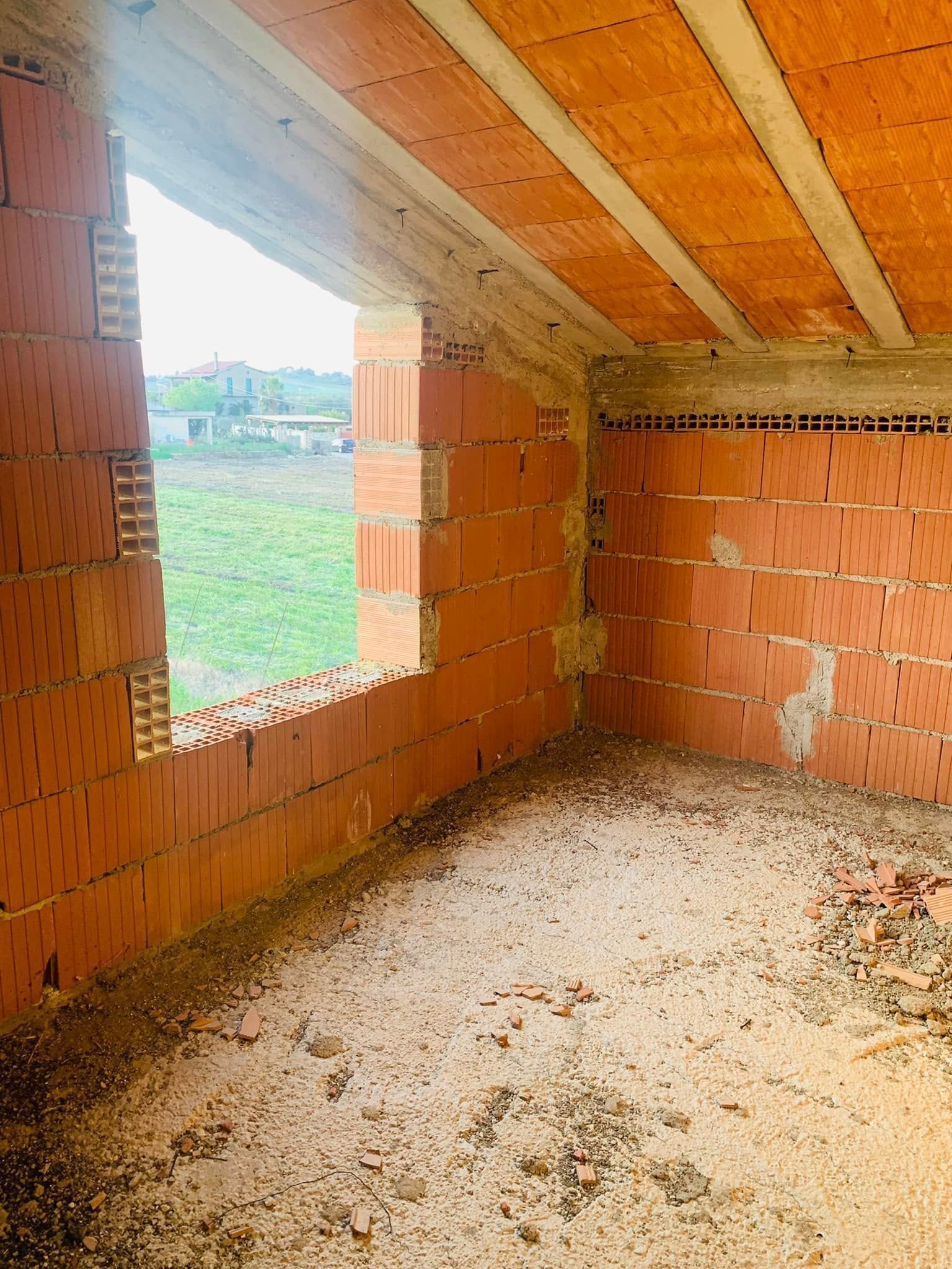Stanza in mattoni non rifinita con soffitto inclinato in legno e una finestra che si apre su un campo erboso.