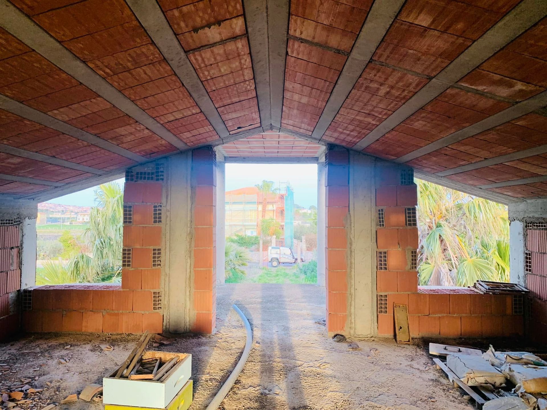 Interno di una casa in mattoni non rifinita con tetto spiovente in legno, porta d'ingresso aperta e finestre che si affacciano sul verde.