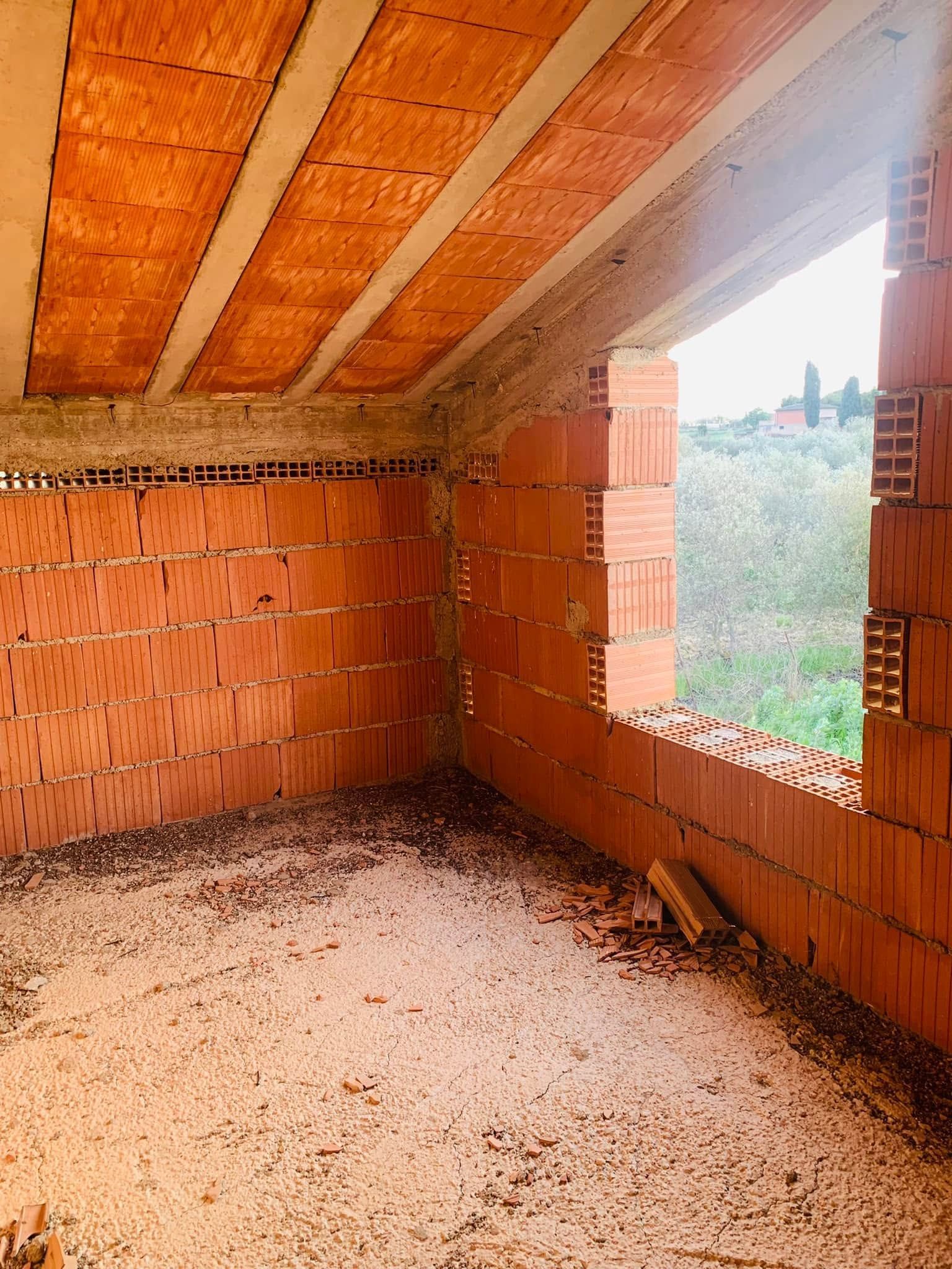 Stanza in mattoni non rifinita con travi in legno a vista sul soffitto e una finestra che si affaccia sul verde.