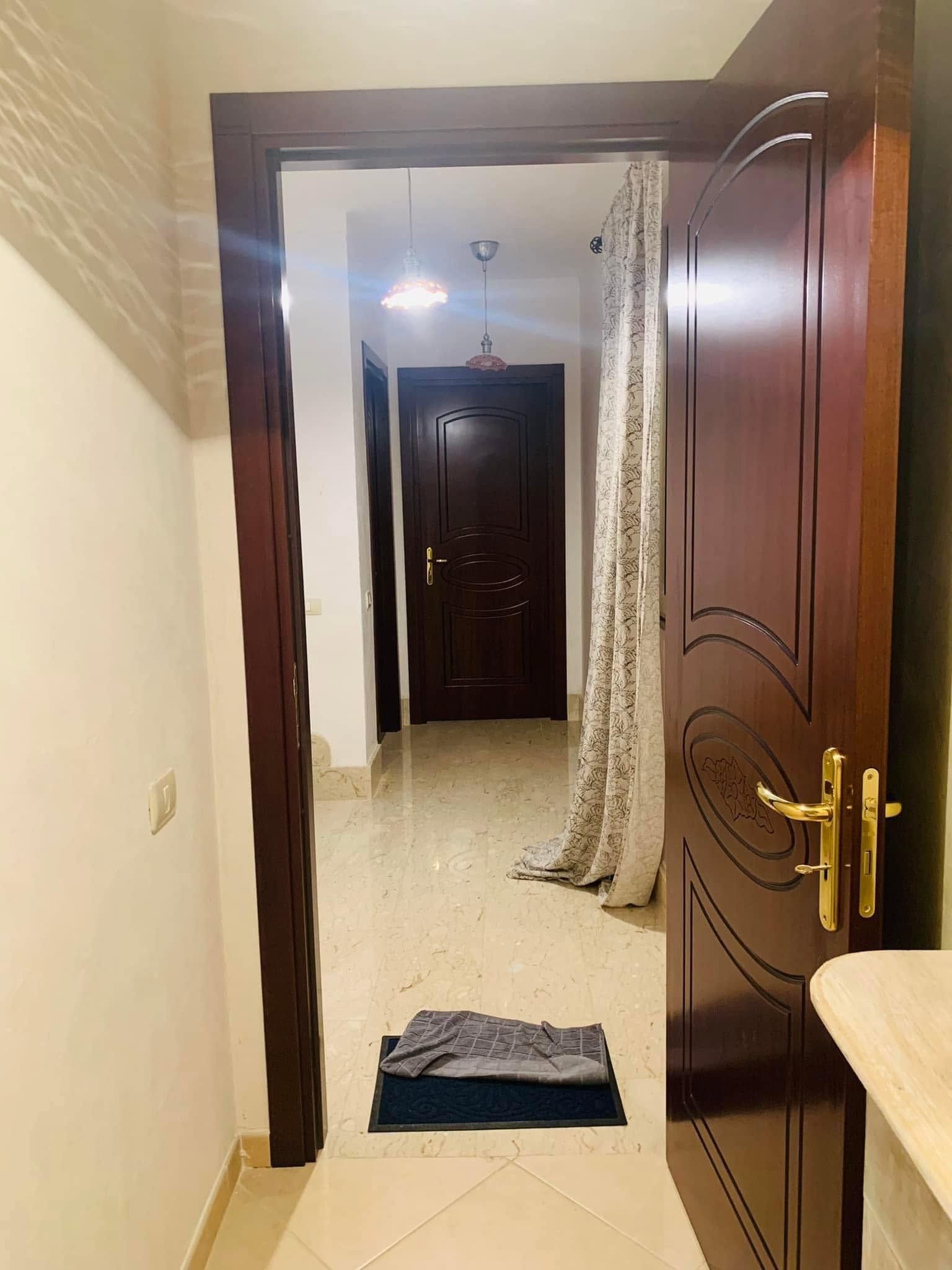 Vista del corridoio attraverso una porta di legno aperta, con un corridoio beige, porte scure e una tenda appesa di fronte.