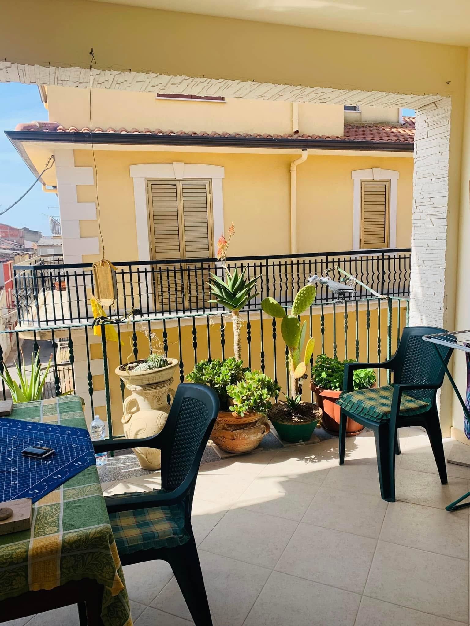 Balcone soleggiato con sedie nere, piante in vaso e un edificio giallo al di là di una ringhiera nera.