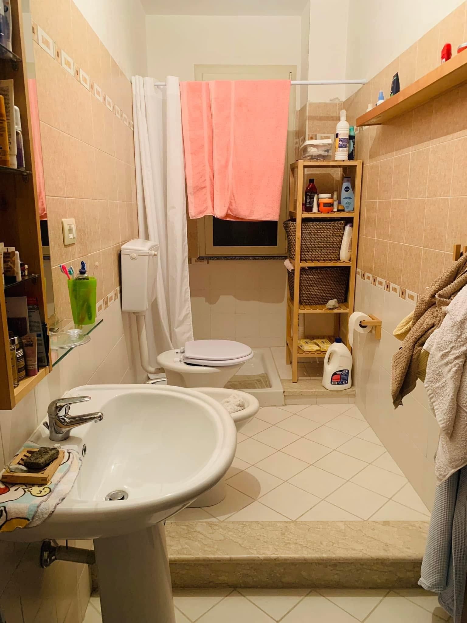 Bagno stretto di colore beige con lavabo, WC, tenda da doccia, mensole e pavimento piastrellato.