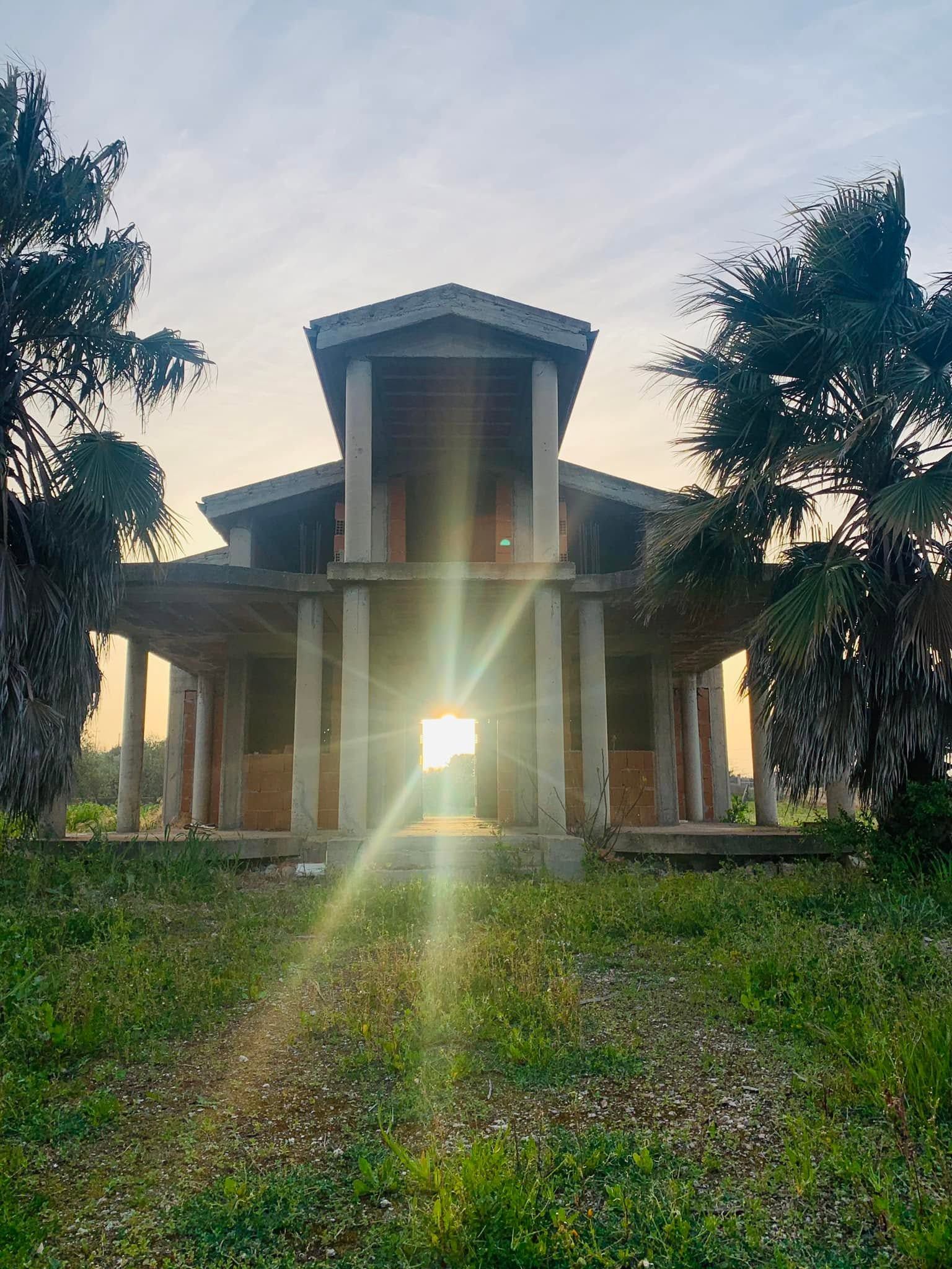 Padiglione illuminato dal sole con colonne e alberi, visto da un sentiero erboso al crepuscolo.