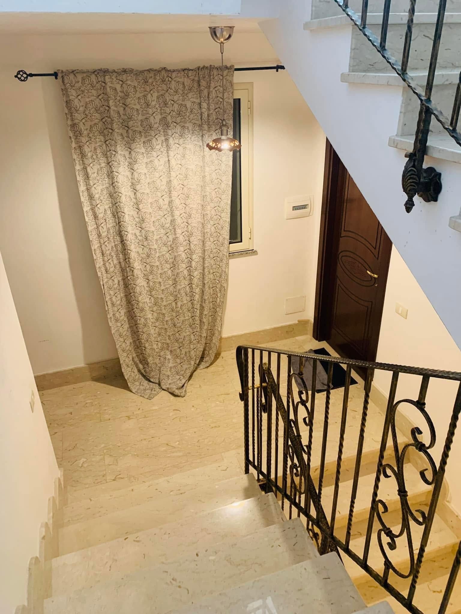 Vano scale interno con pareti bianche, pavimento beige, porta in legno scuro e tenda a motivi geometrici appesa a una finestra.