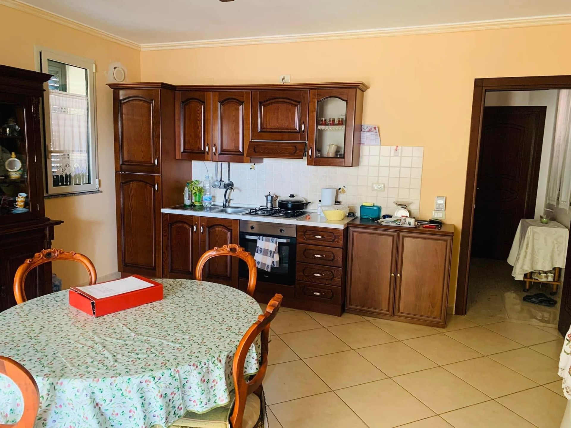 Cucina e zona pranzo con mobili in legno, pavimento piastrellato, tavolo rotondo e porta che conduce a un'altra stanza.