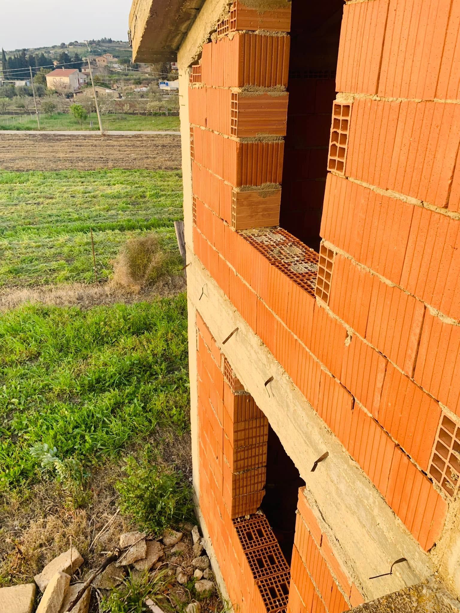 Muro di mattoni arancioni parzialmente costruito con aperture finestrate che si affacciano su un campo verde.