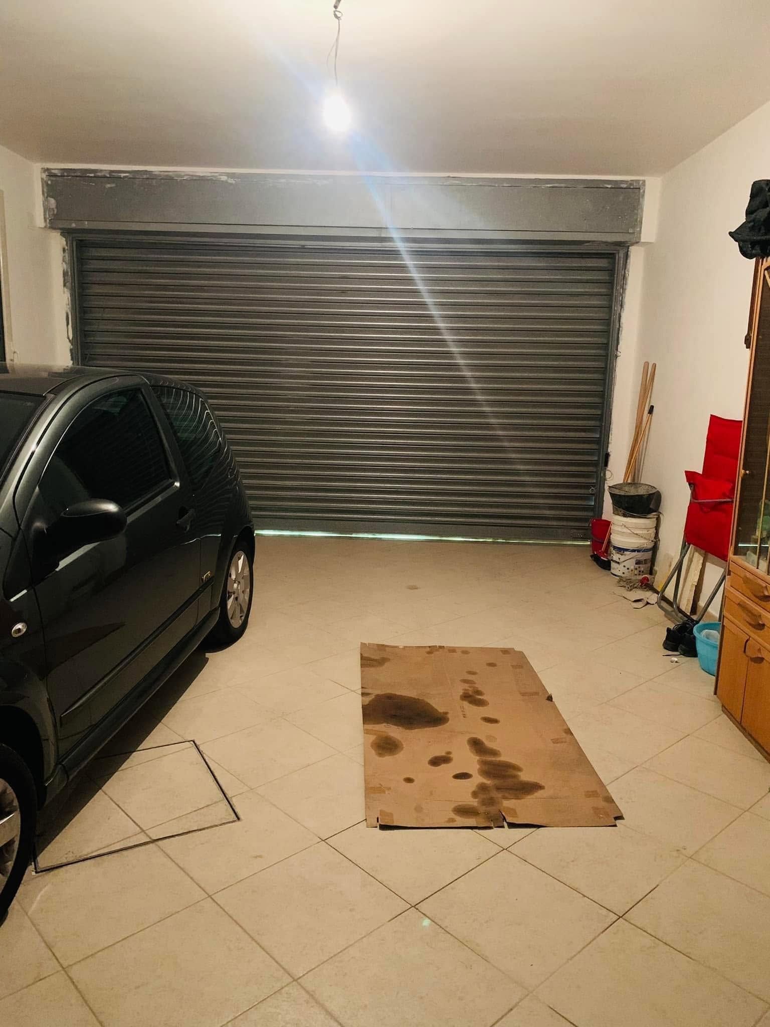 Interno di un garage con un'auto nera, porta metallica chiusa, pavimento piastrellato e un foglio di cartone sul pavimento.