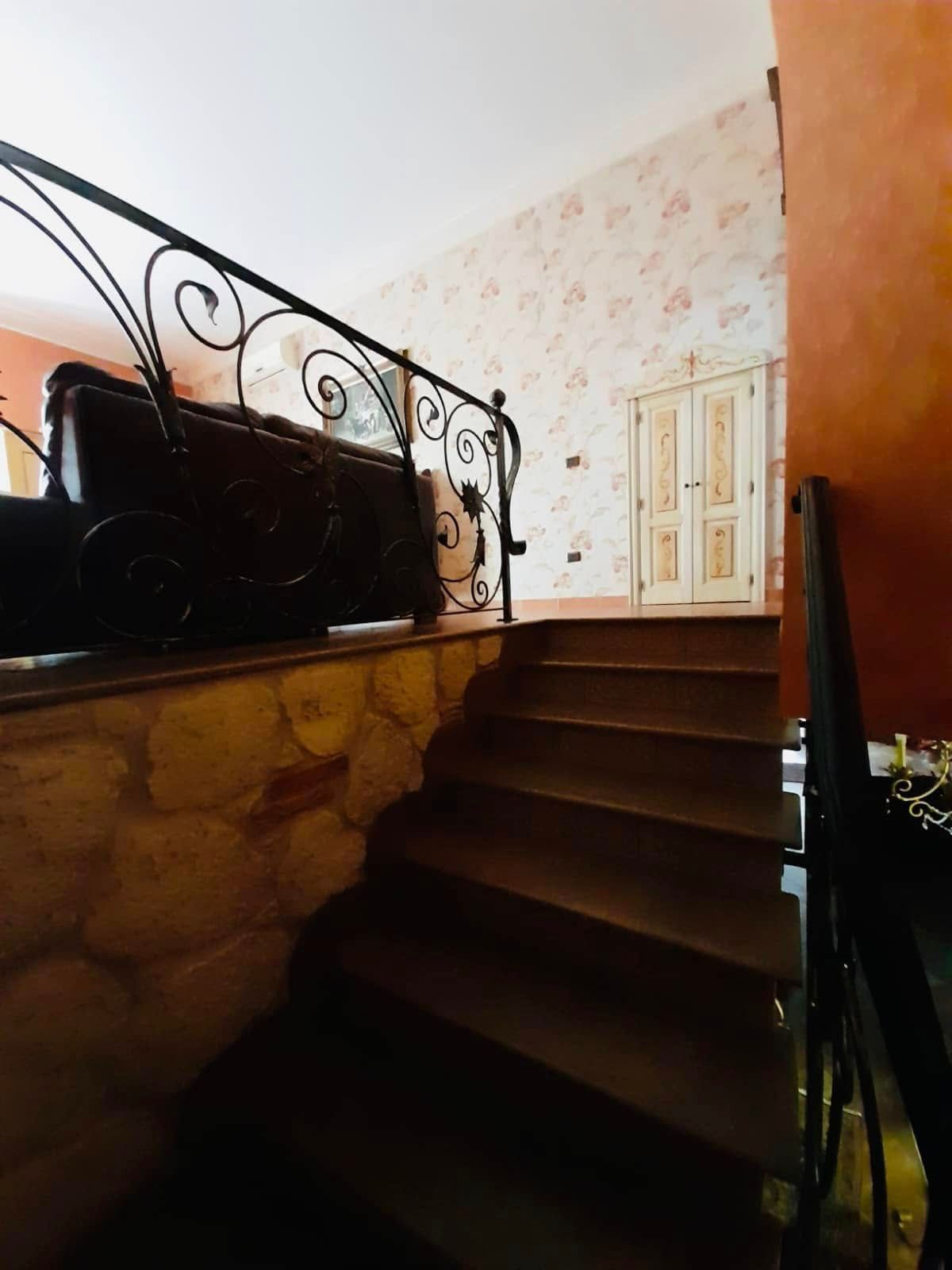 Scala interna con gradini scuri, ringhiera in ferro battuto, parete in pietra e carta da parati floreale.