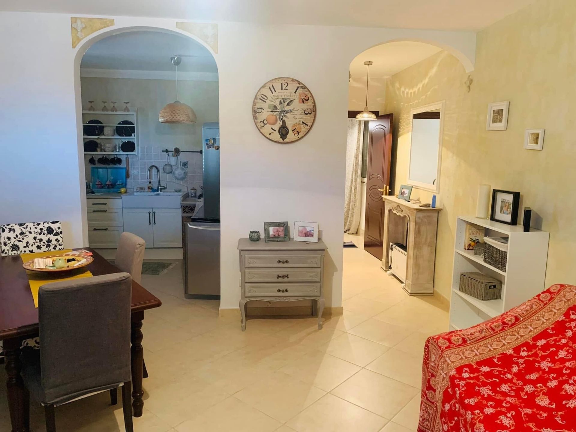 Luminoso soggiorno con archi, tavolo da pranzo, orologio e poltrona con motivo rosso; cucina visibile sul retro.
