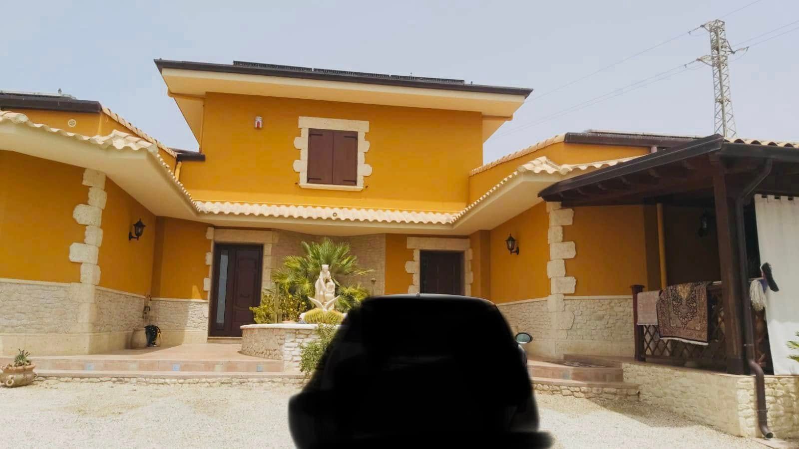 Casa gialla con tetto di tegole e veranda ombreggiata, vista dal cortile anteriore.