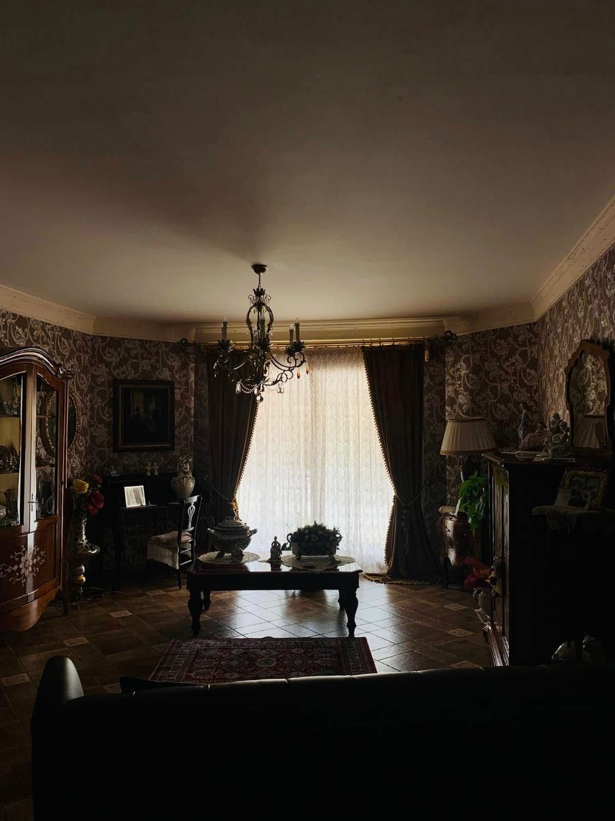 Soggiorno sfarzoso e poco illuminato con carta da parati decorata, lampadario e finestra luminosa con tende pesanti.