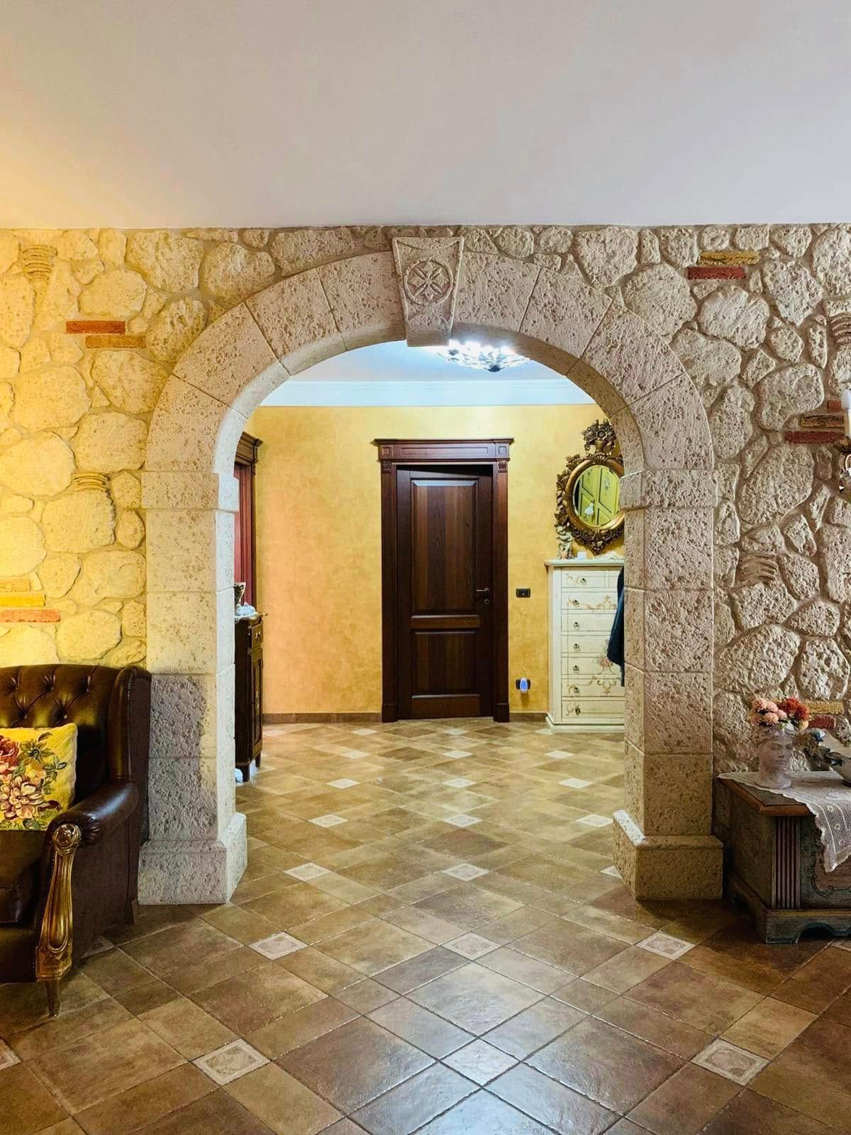 Corridoio ad arco in pietra con pavimento piastrellato, porta in legno scuro e posti a sedere in un interno illuminato da una luce calda.