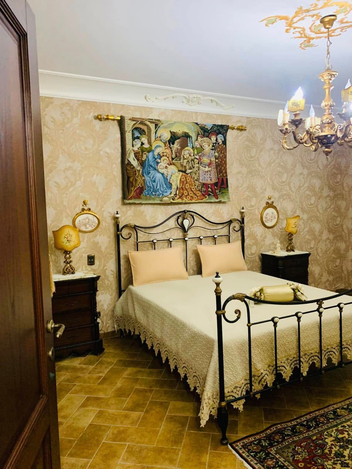 Elegante camera da letto con letto in ferro battuto decorato, pareti beige, lampadario e tappeto a motivi geometrici.