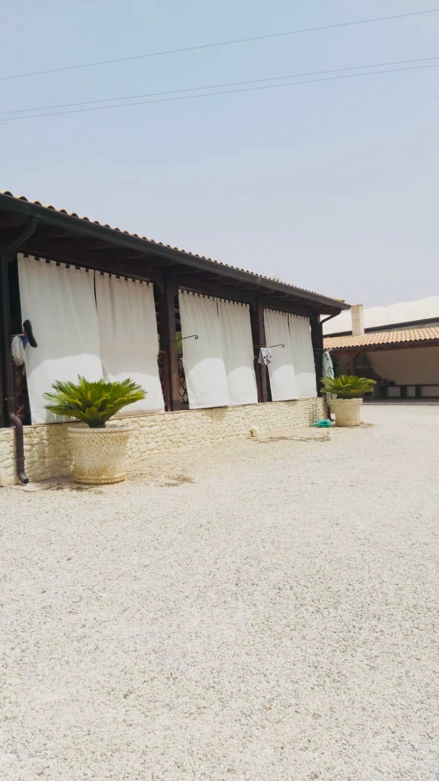 Cortile di ghiaia accanto a un lungo edificio bianco con tetto scuro e piante in vaso sotto un cielo limpido