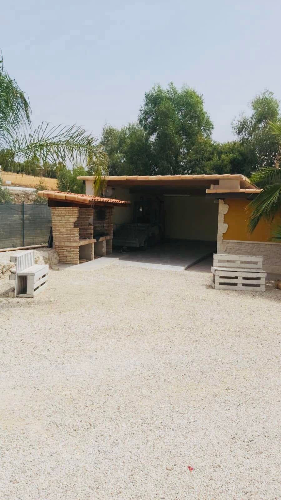 Vialetto di ghiaia che conduce a un posto auto coperto e ombreggiato, con pallet impilati e alberi sullo sfondo.