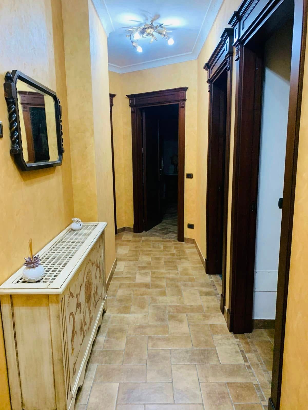 Stretto corridoio con pavimento piastrellato beige, porte in legno scuro, specchio a parete e mobiletto con termosifone.
