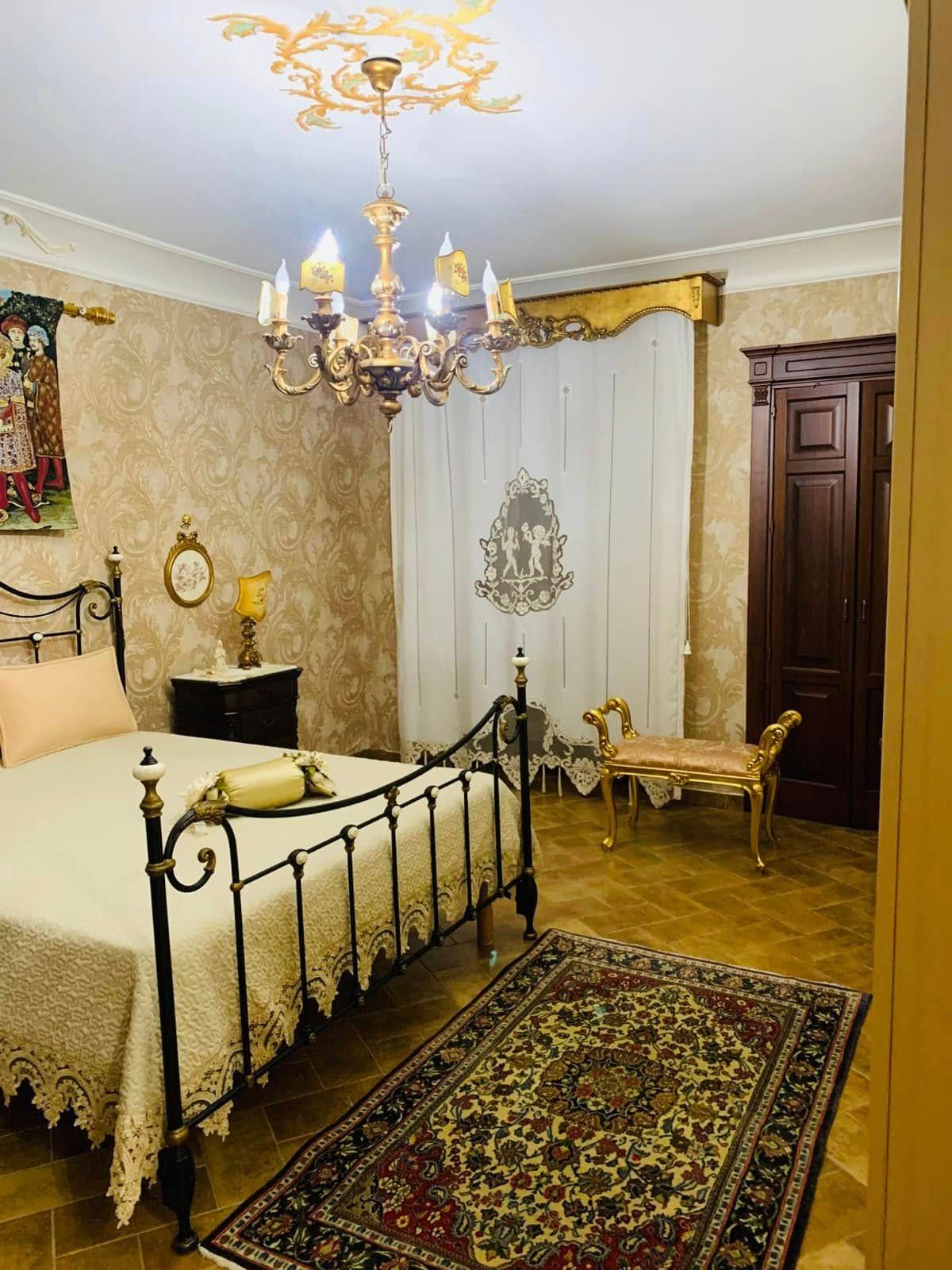 Elegante camera da letto in stile vintage con letto decorato, lampadario, tappeto a motivi geometrici e tende bianche drappeggiate.