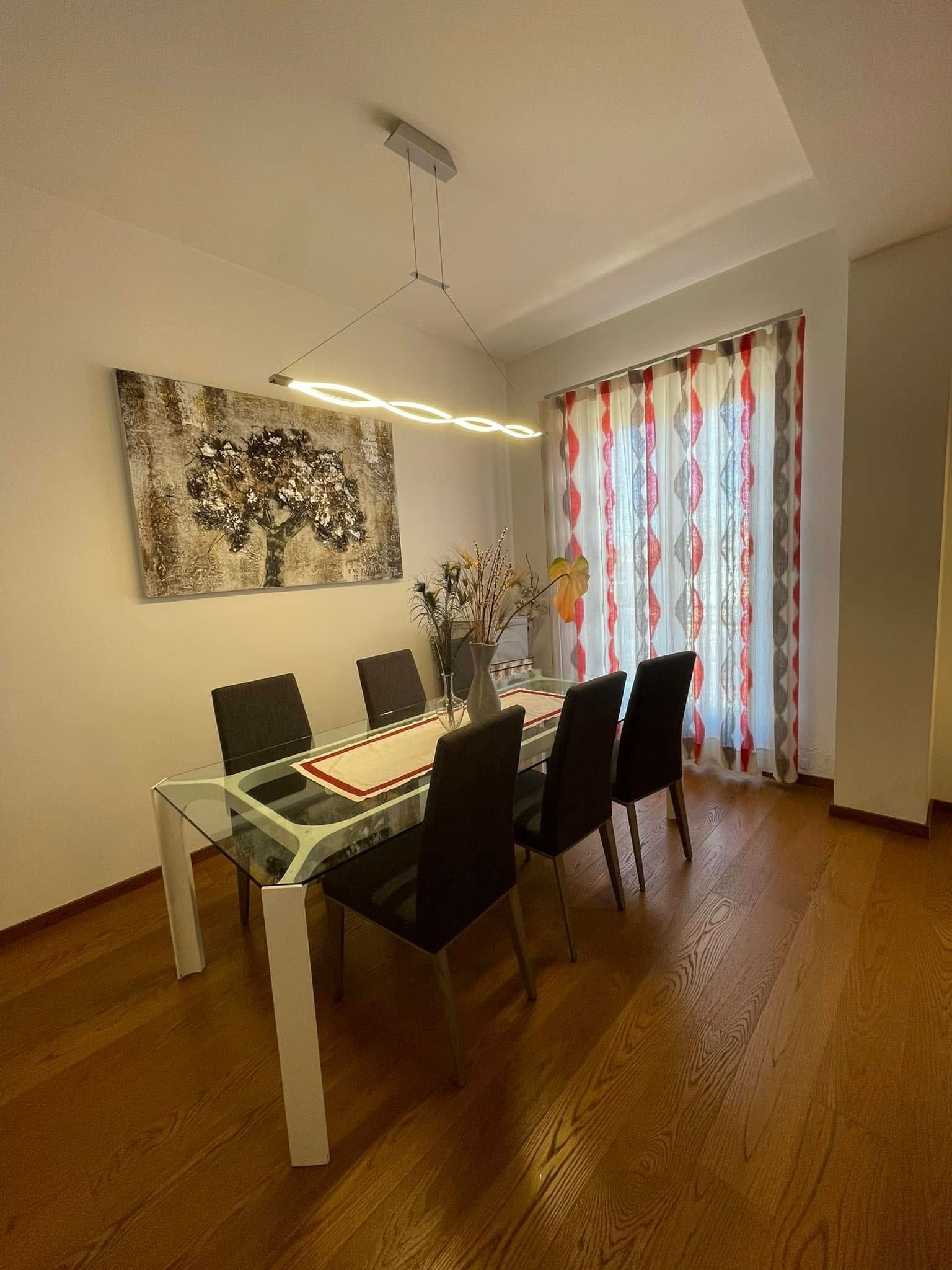 Sala da pranzo moderna con tavolo in vetro, sedie nere, opere d'arte alle pareti e tende trasparenti con motivi decorativi.