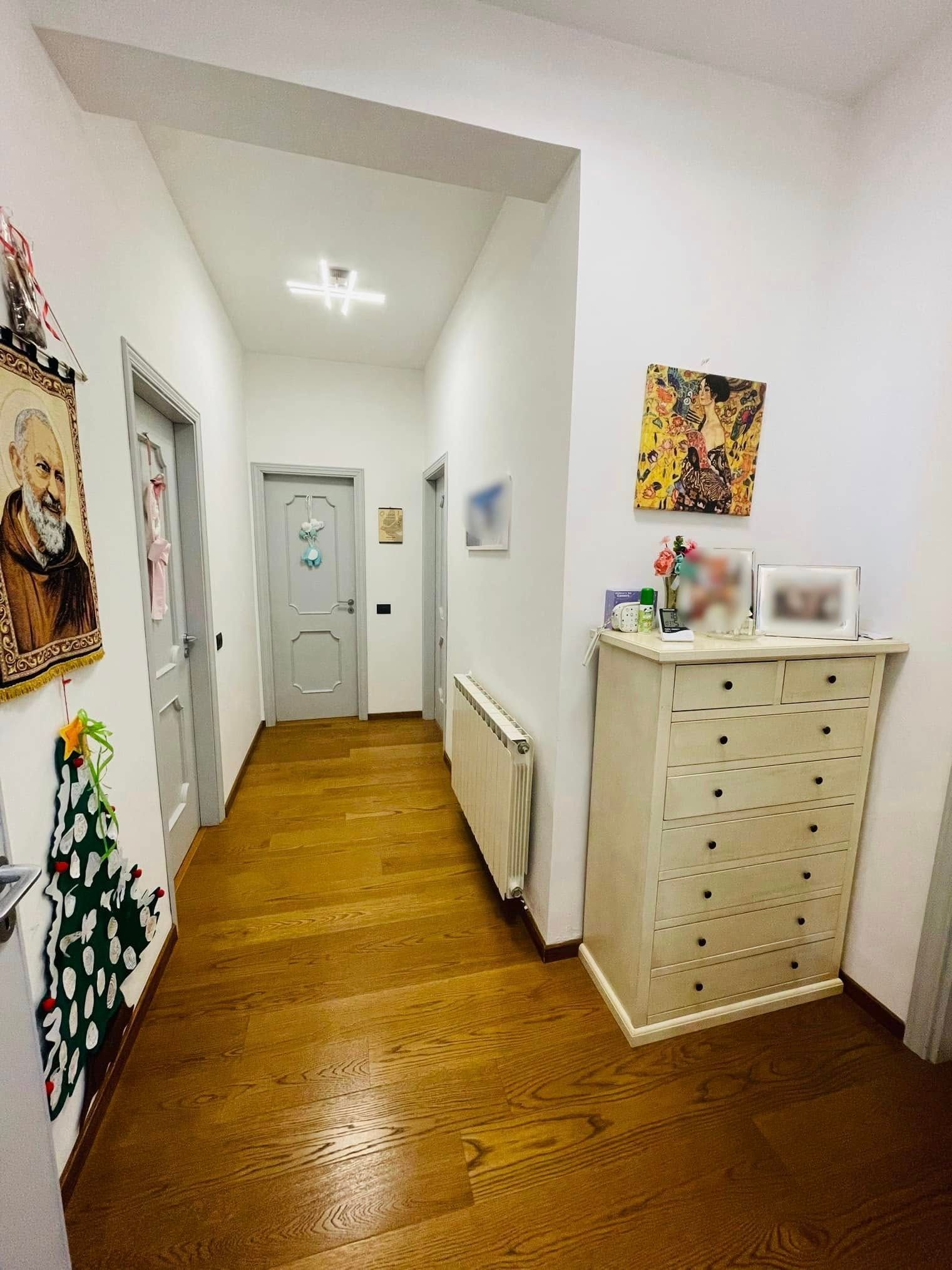 Stretto corridoio bianco con pavimento in legno, termosifone, quadri alle pareti e una cassettiera bianca in fondo.