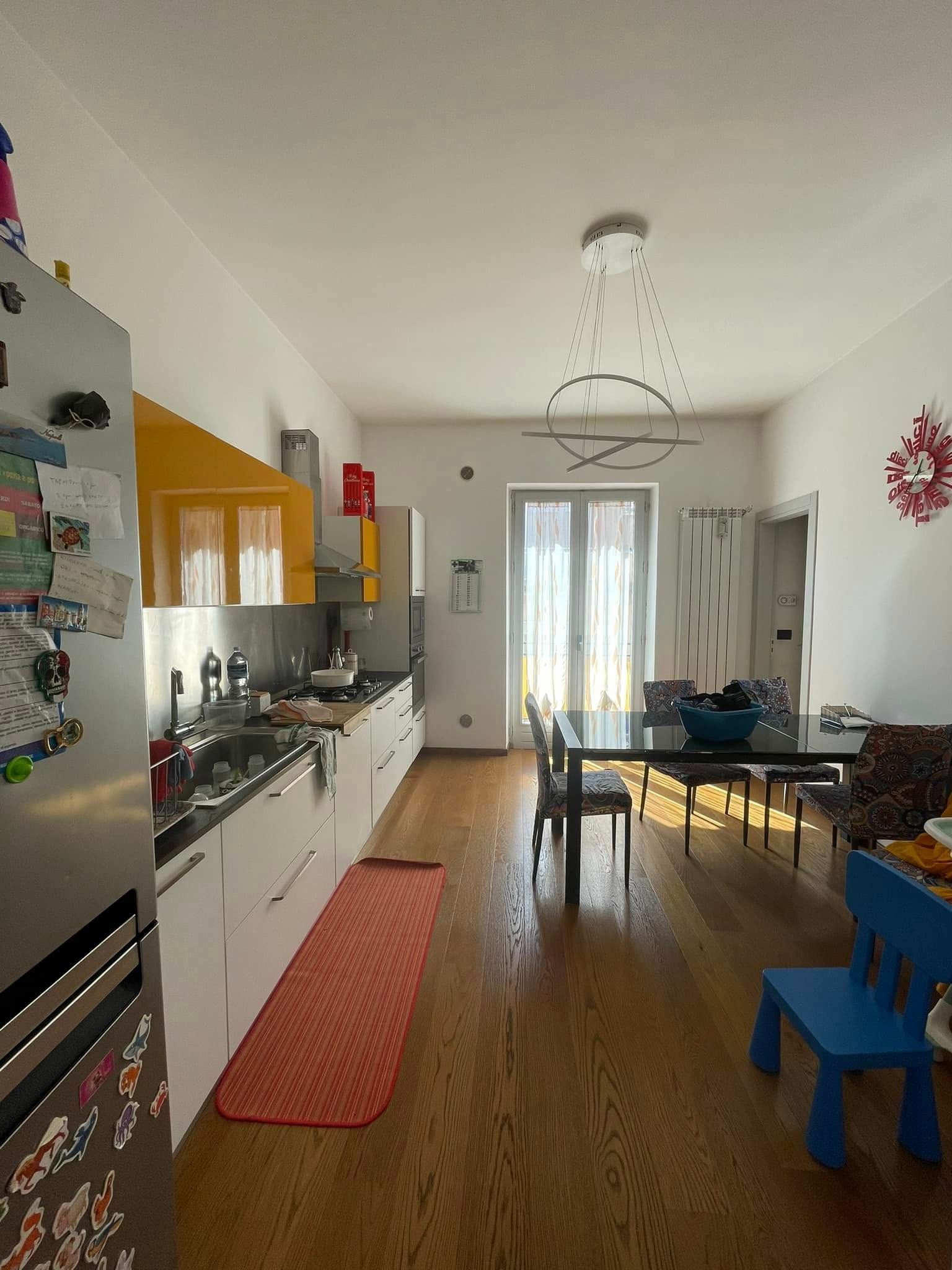 Cucina e zona pranzo luminose con mobili bianchi, pavimento in legno, tavolo vicino alla finestra e sedia blu in primo piano.