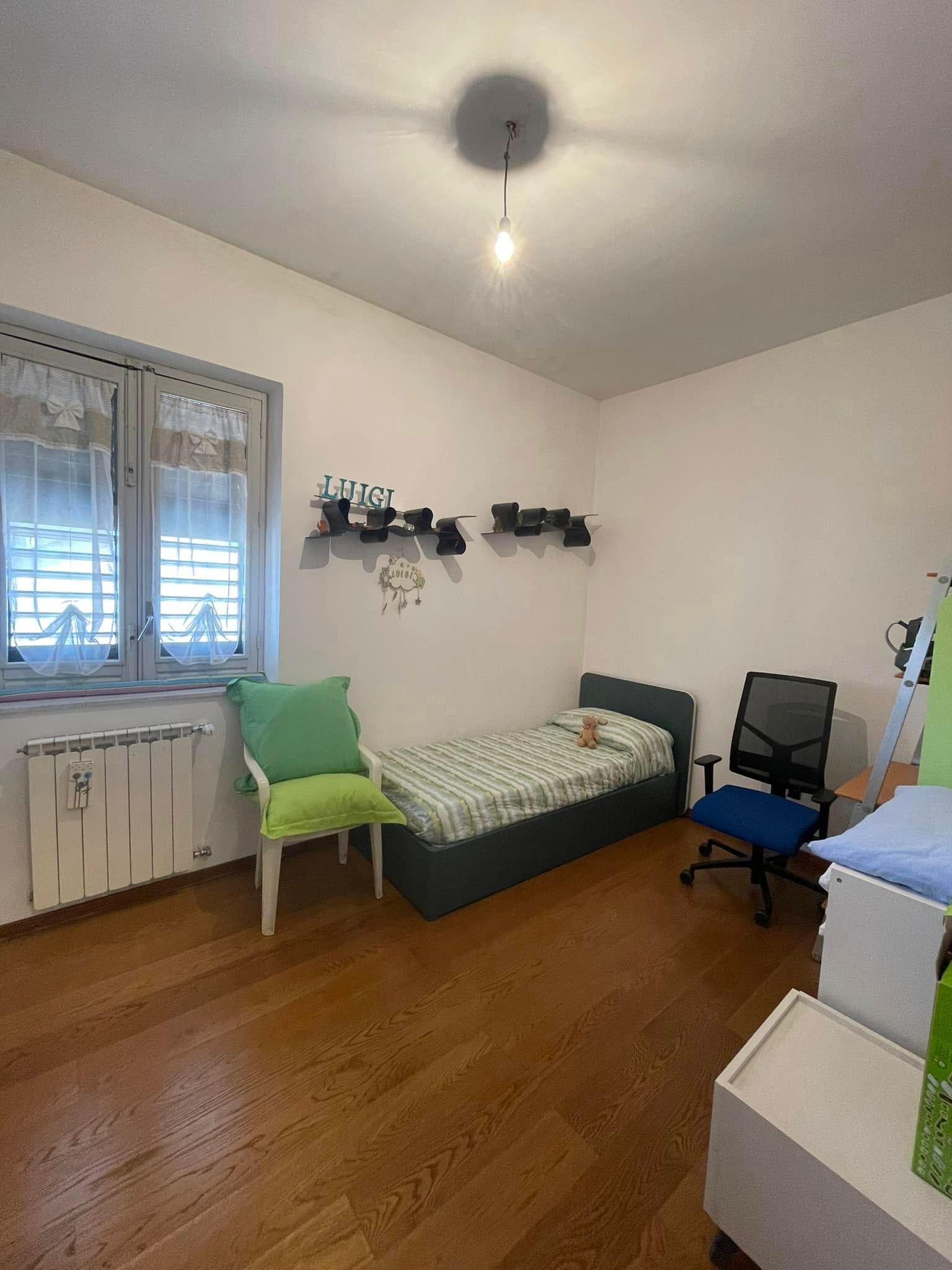 Piccola camera da letto con due letti singoli, sedia da scrivania, finestra e termosifone bianco, illuminato da una lampadina a soffitto.