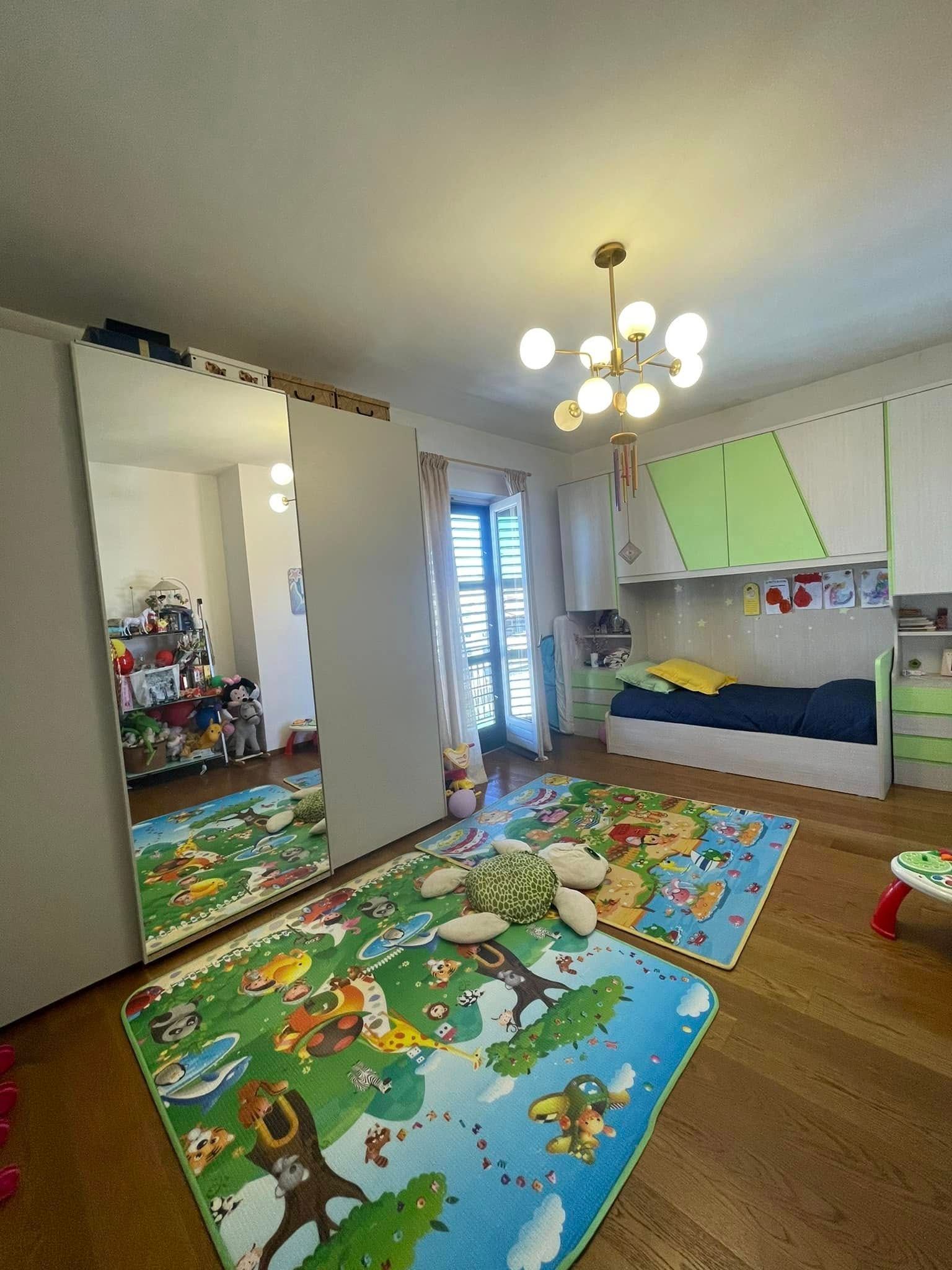 Una cameretta colorata per bambini con letto, tappeto cosparso di giocattoli, armadio con specchio e lampadario.