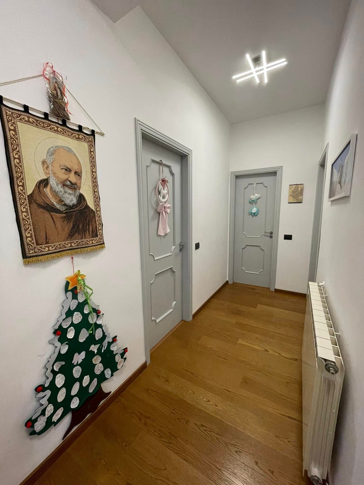 Stretto corridoio con pareti bianche, pavimento in legno, porte decorate, un ritratto sulla sinistra e una piccola sagoma di albero di Natale.