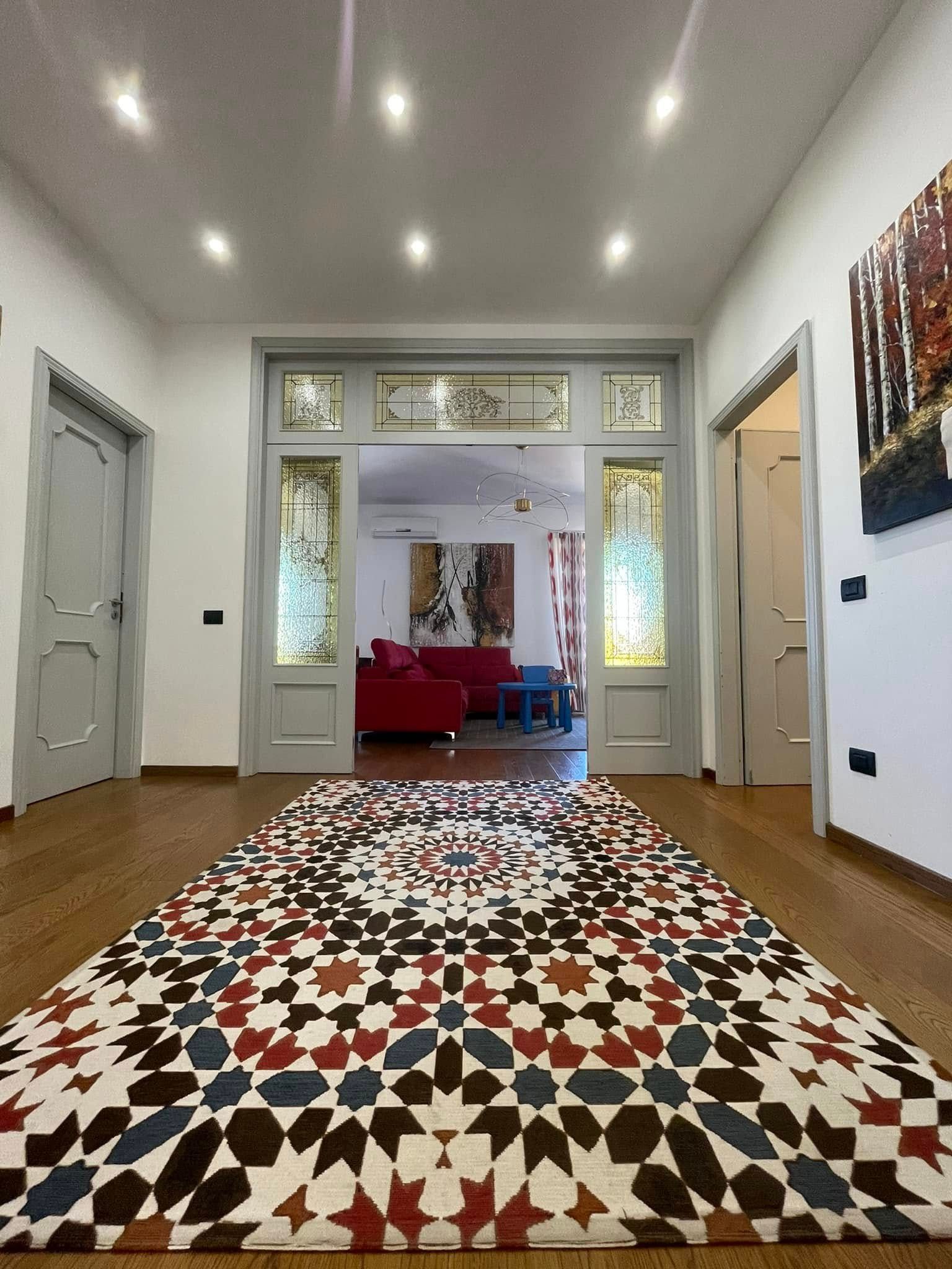 Luminoso corridoio con tappeto a motivi geometrici, pareti bianche, faretti a incasso e sedute rosse visibili attraverso le porte aperte.