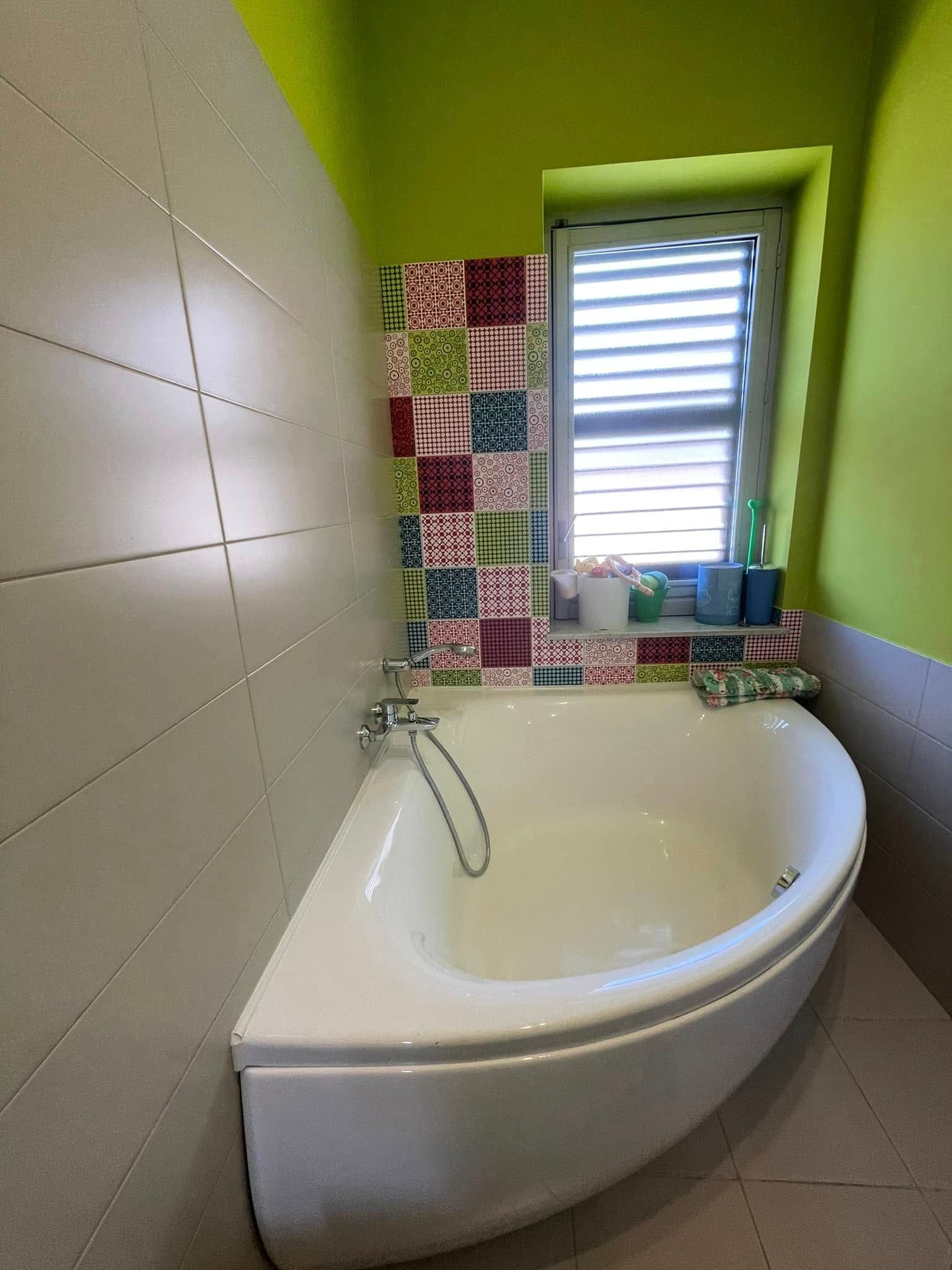 Piccolo bagno con vasca angolare, pareti verdi, piastrelle decorate e una finestra sopra la vasca.