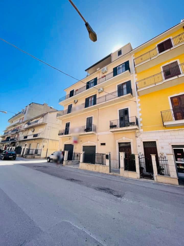 Una strada soleggiata con palazzi dai colori pastello, balconi e auto parcheggiate sotto un cielo azzurro.