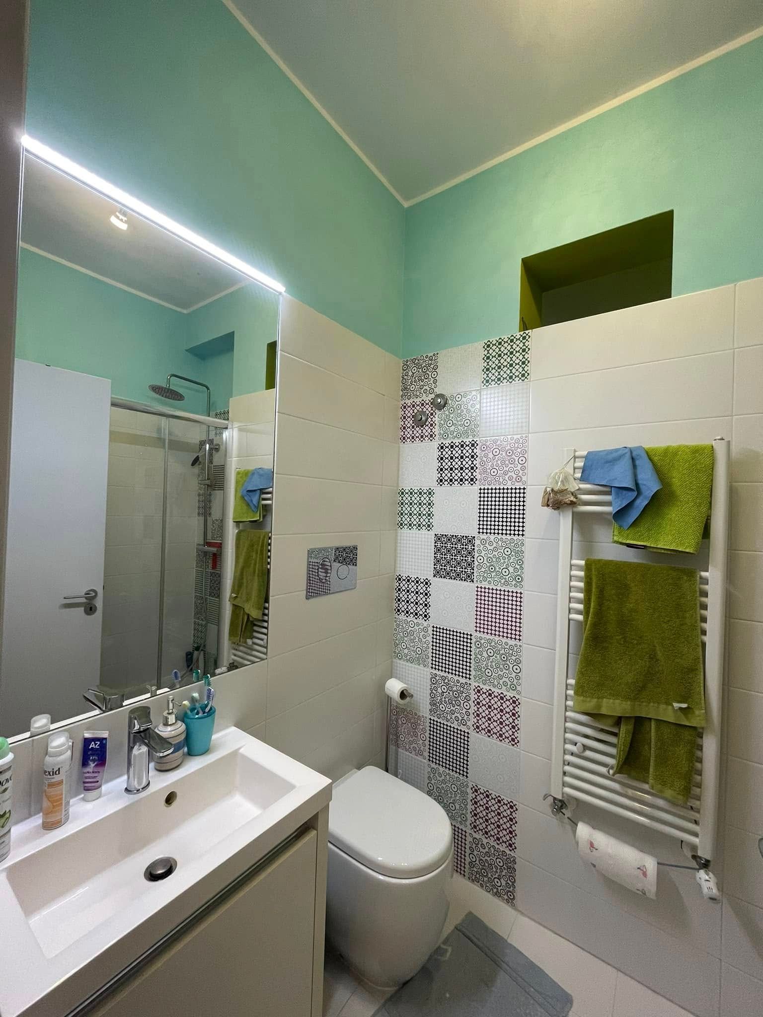 Bagno compatto con lavabo, specchio, WC, doccia, pareti color verde acqua e piastrelle decorative decorate.
