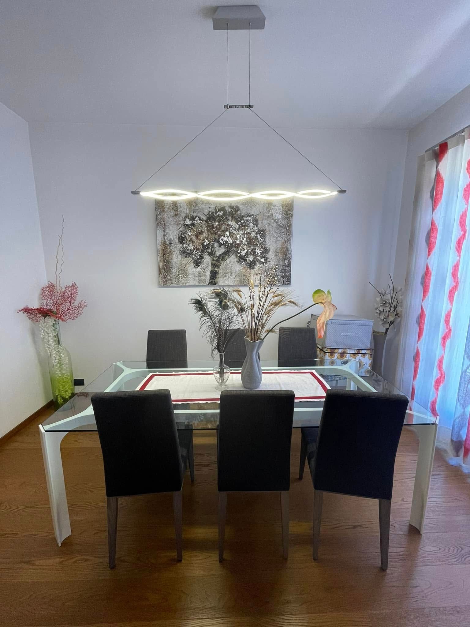 Sala da pranzo moderna con tavolo apparecchiato, sedie nere, lampada a sospensione e tende vicino a una finestra.