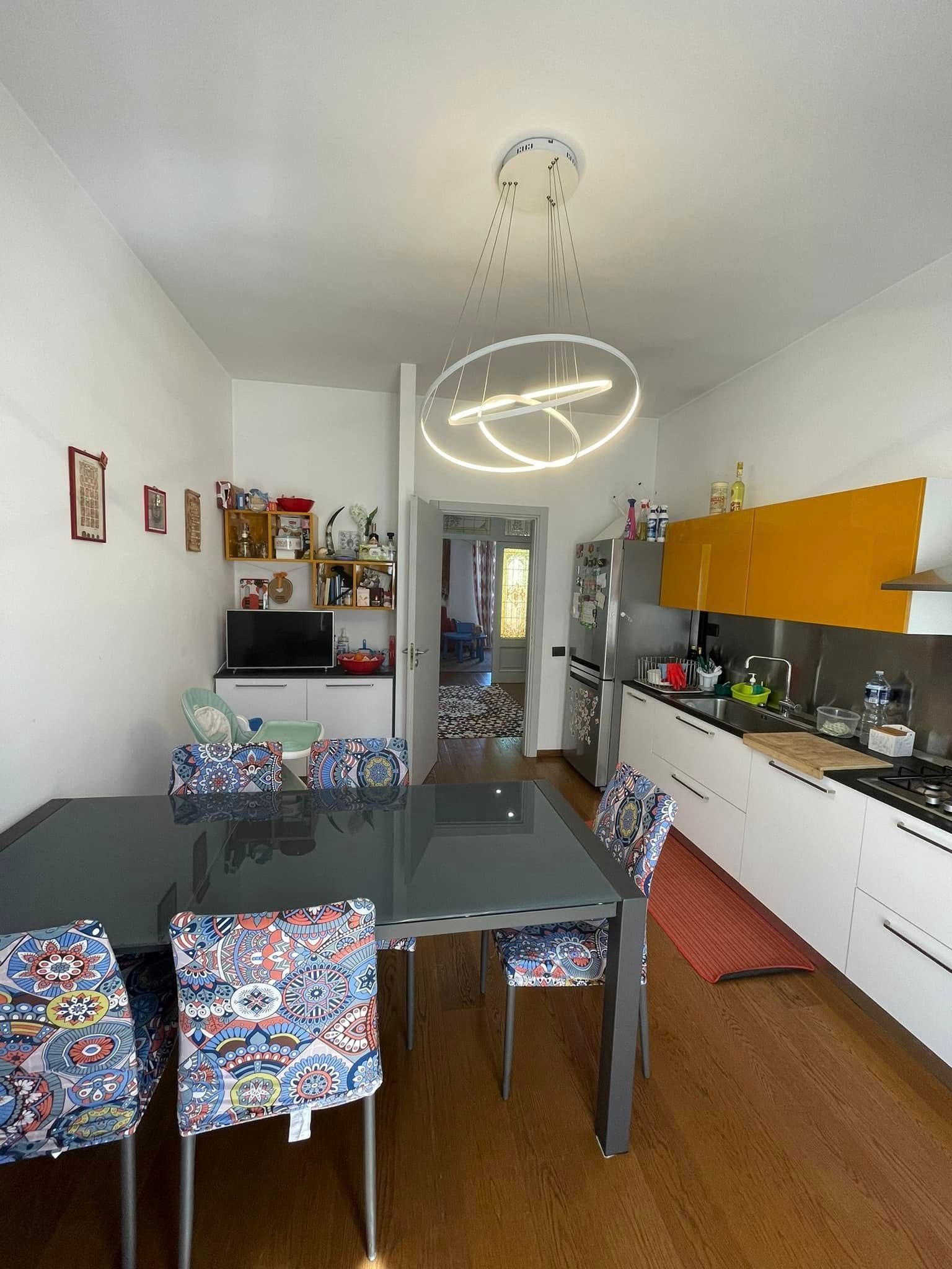 Cucina e zona pranzo moderne con sedie colorate, mobili bianchi, paraschizzi giallo e lampada ad anello sospesa.