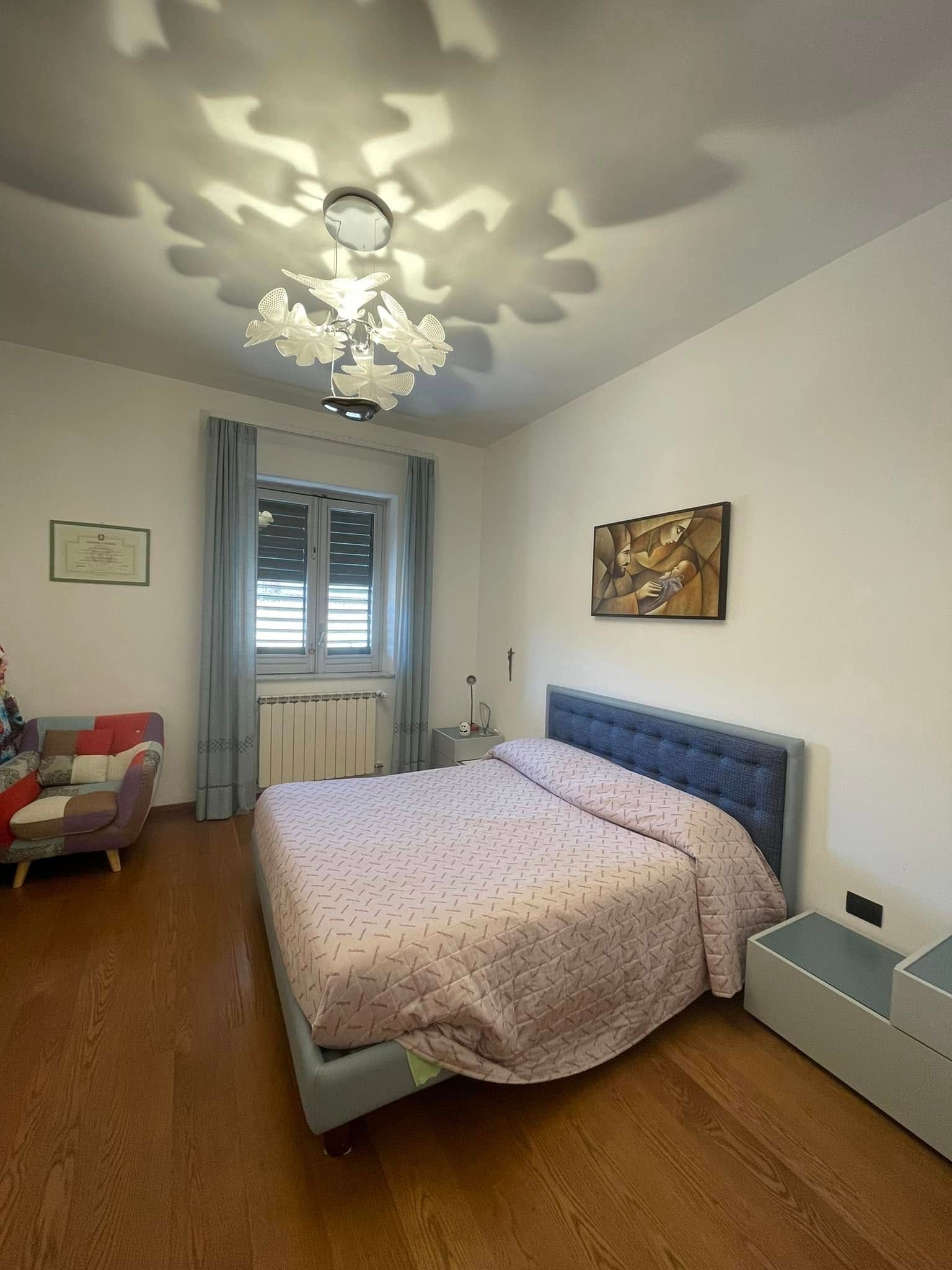 Camera da letto luminosa con letto, finestra, pavimento in legno e lampadario a soffitto.