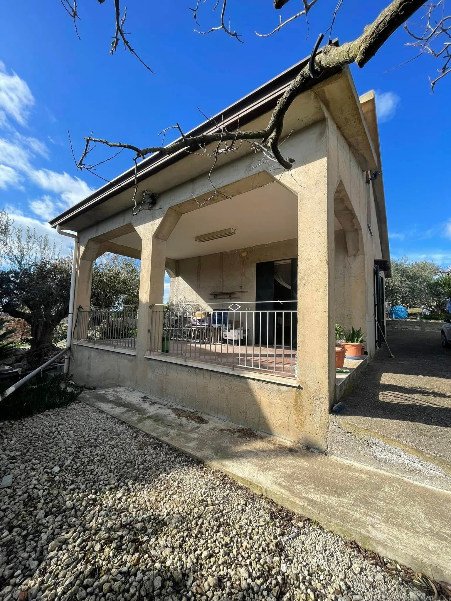 Piccolo padiglione beige con tetto spiovente e portico anteriore in un cortile di ghiaia sotto un cielo azzurro limpido.