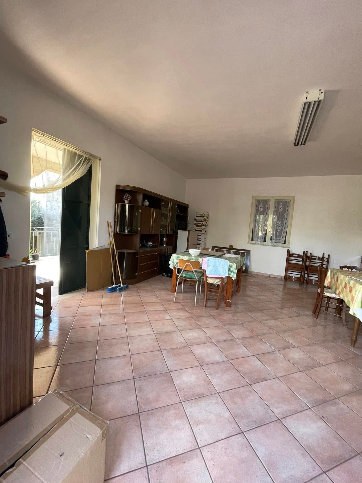 Ampia sala da pranzo piastrellata con tavolo in legno, credenza, sedie e porta aperta su una luminosa area esterna.