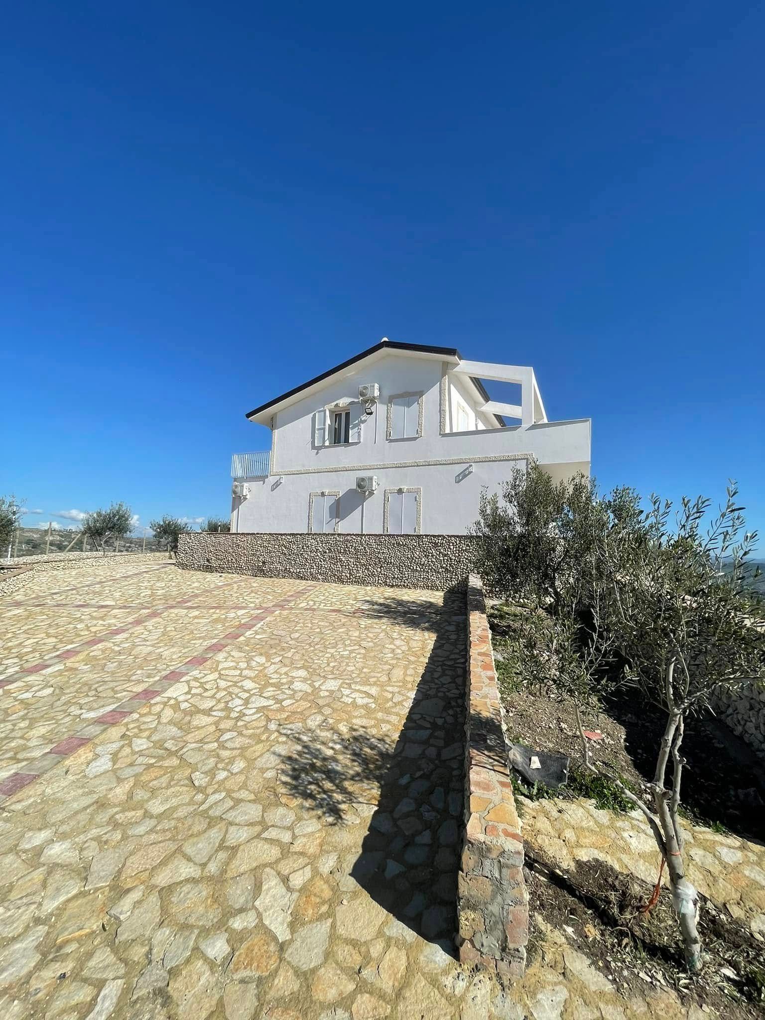 Casa bianca su una collina soleggiata con vialetto in pietra e cielo azzurro.