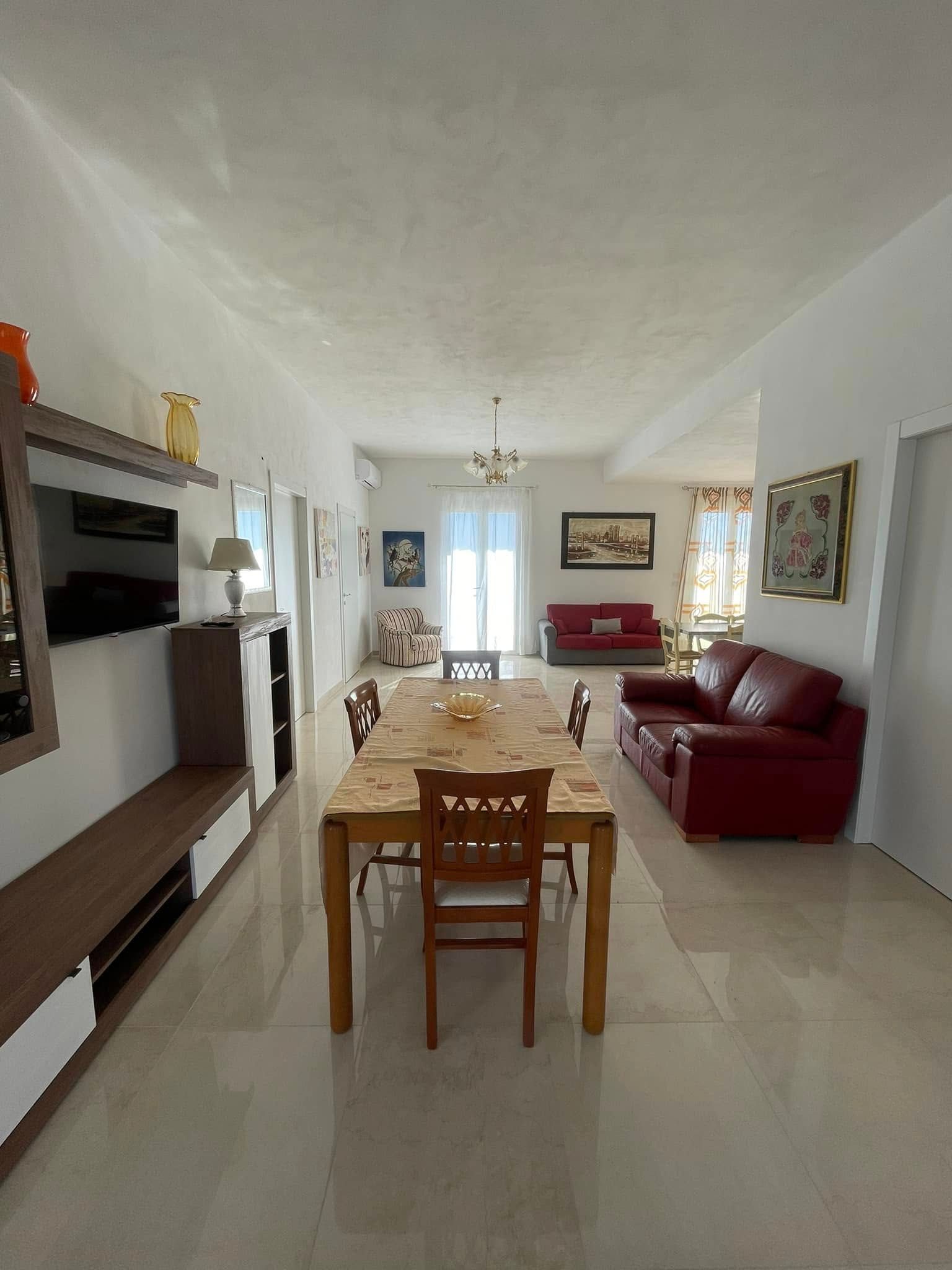 Luminoso soggiorno con divano rosso, tavolo da pranzo in legno, TV a parete e porta aperta su un balcone.