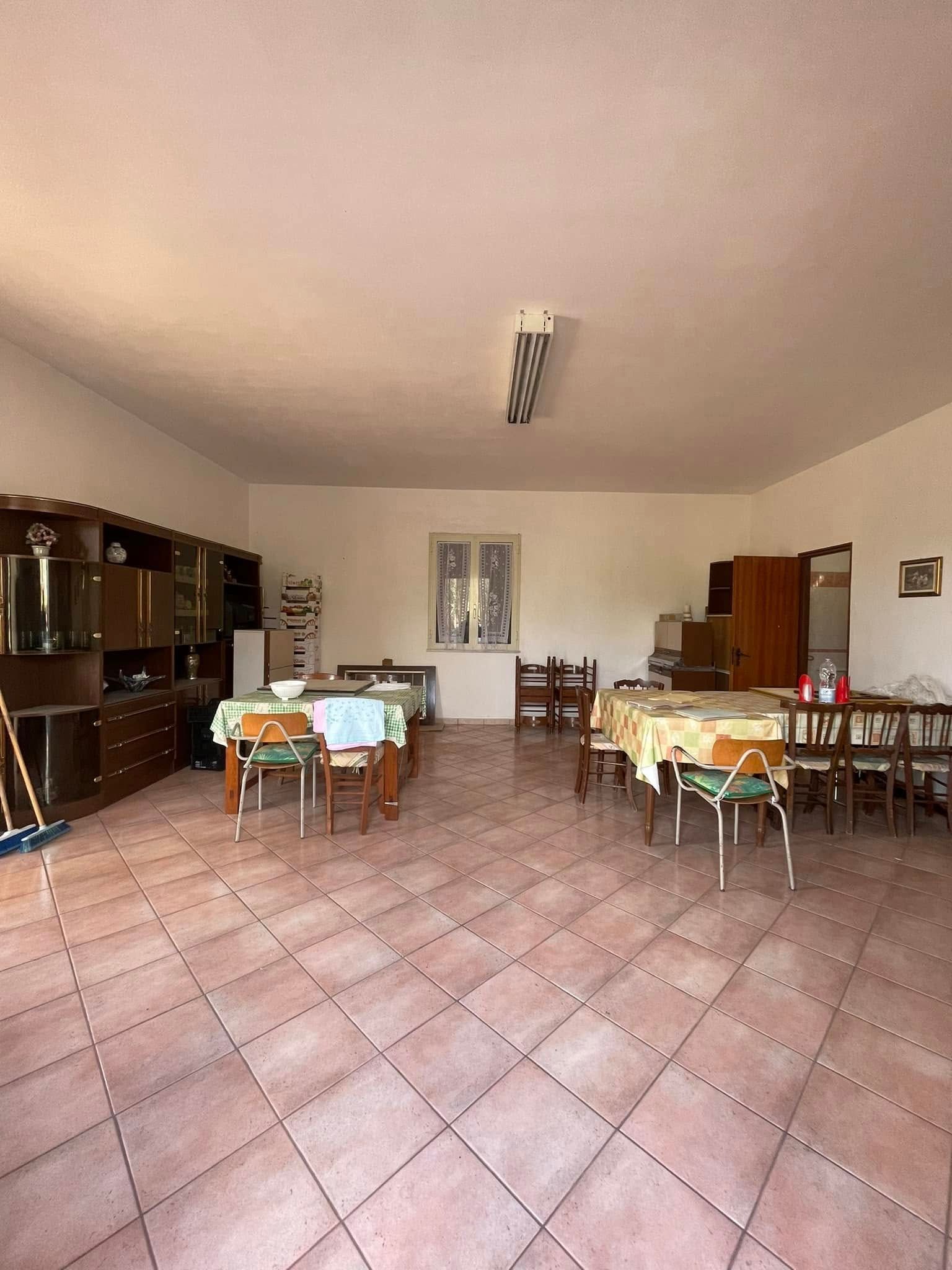 Sala da pranzo con pavimento piastrellato, tavoli e sedie in legno e armadietti lungo le pareti.