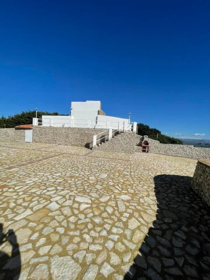 Edificio bianco su una collina lastricata in pietra sotto un cielo azzurro limpido, con una piccola persona seduta sulla destra.