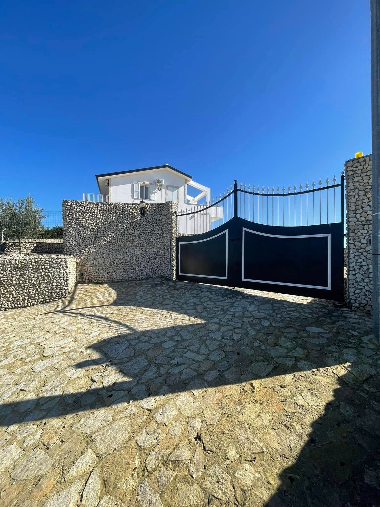 Un vialetto in pietra conduce a un cancello in metallo nero e a una casa bianca sotto un cielo azzurro e limpido.