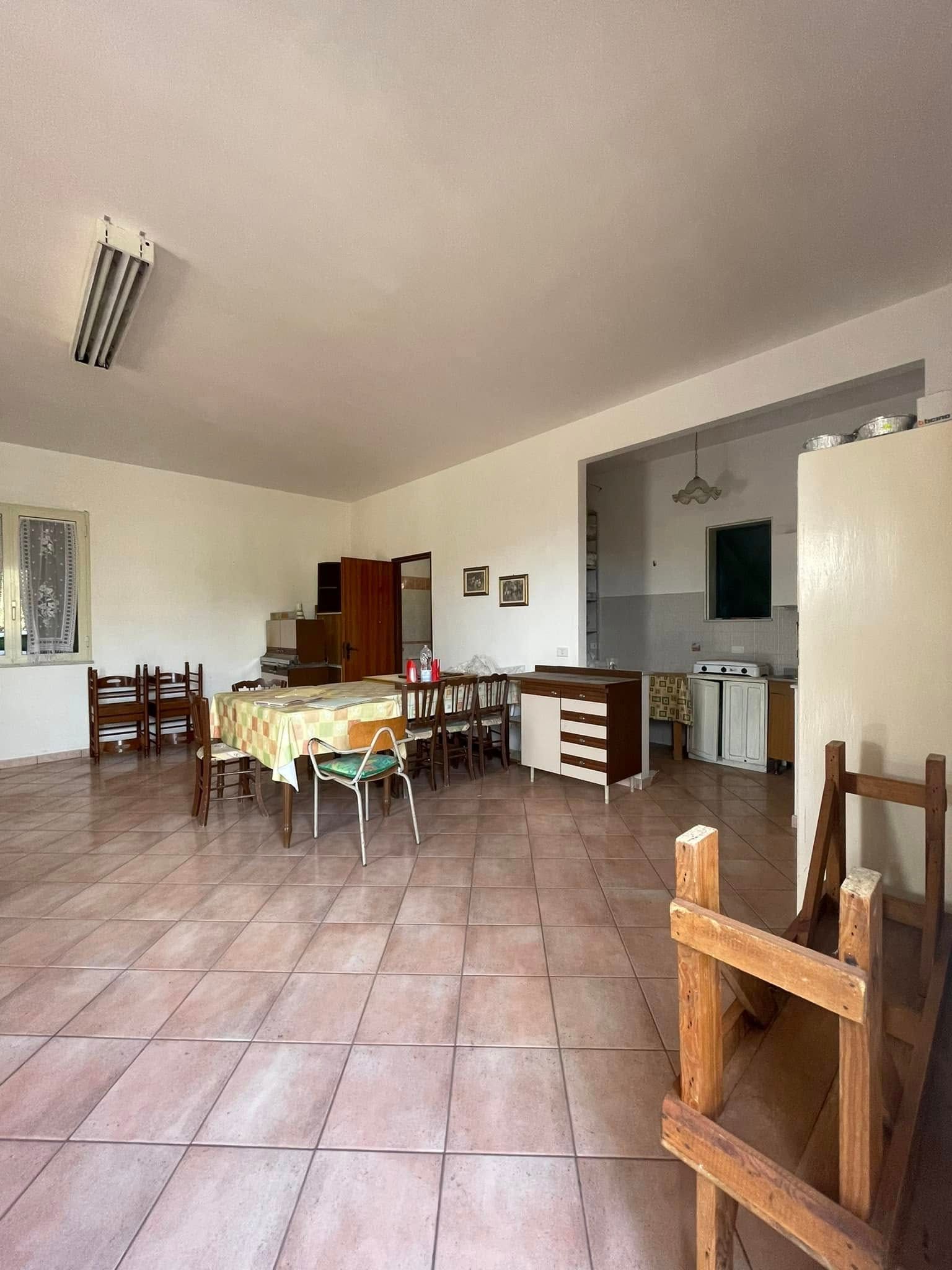 Sala da pranzo vuota con tavoli e sedie in legno, pavimento piastrellato e pareti bianche illuminate da luci intense sul soffitto.