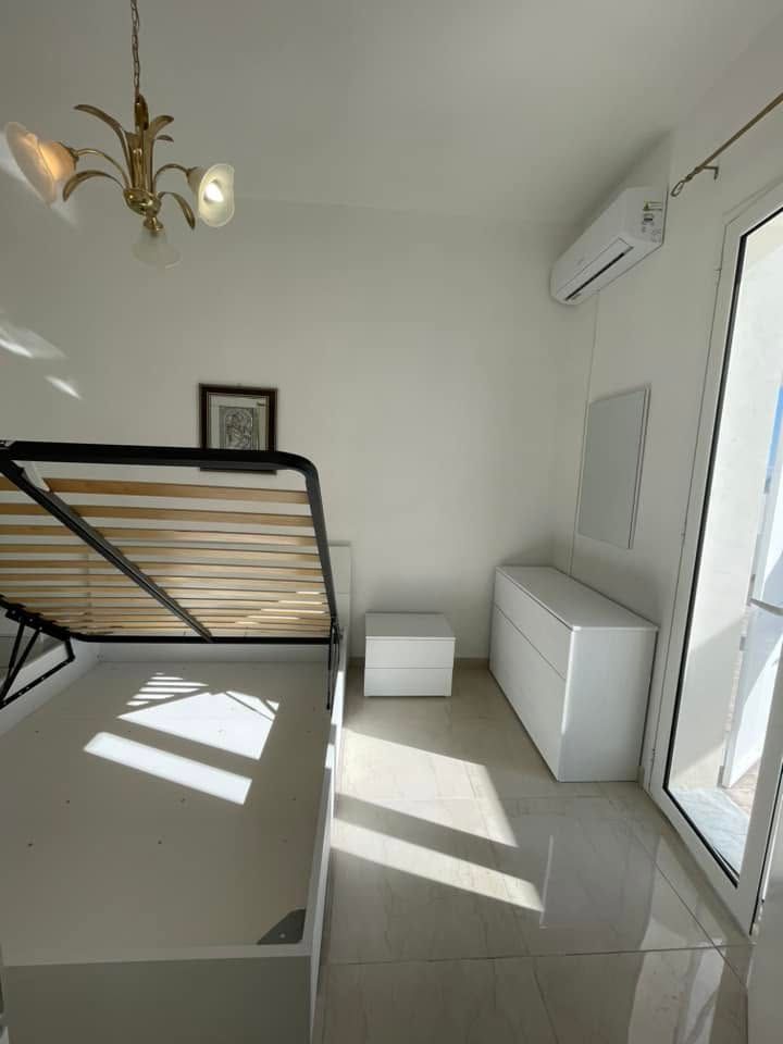 Luminosa camera da letto minimalista con letto, armadio bianco, pavimento piastrellato e luce naturale che filtra attraverso una porta a vetri.