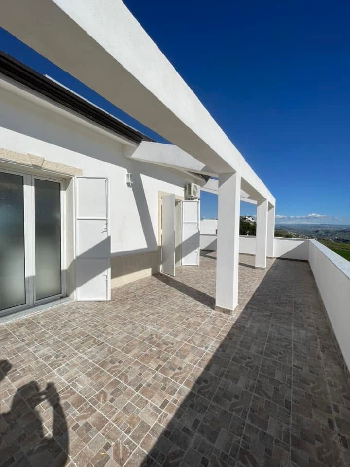Terrazza panoramica soleggiata e bianca con pavimento piastrellato, travi a pergola e vista sull'oceano sotto un cielo azzurro limpido.