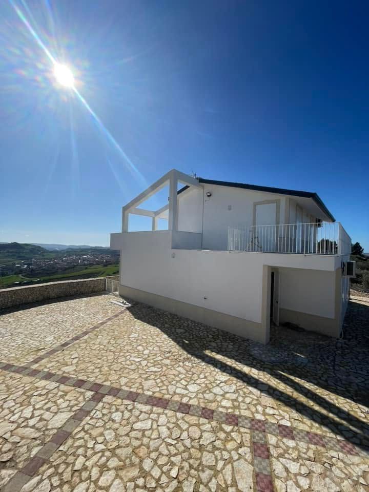 Casa bianca con terrazza sul tetto, situata su una collina soleggiata e visibile da un vialetto lastricato in pietra.