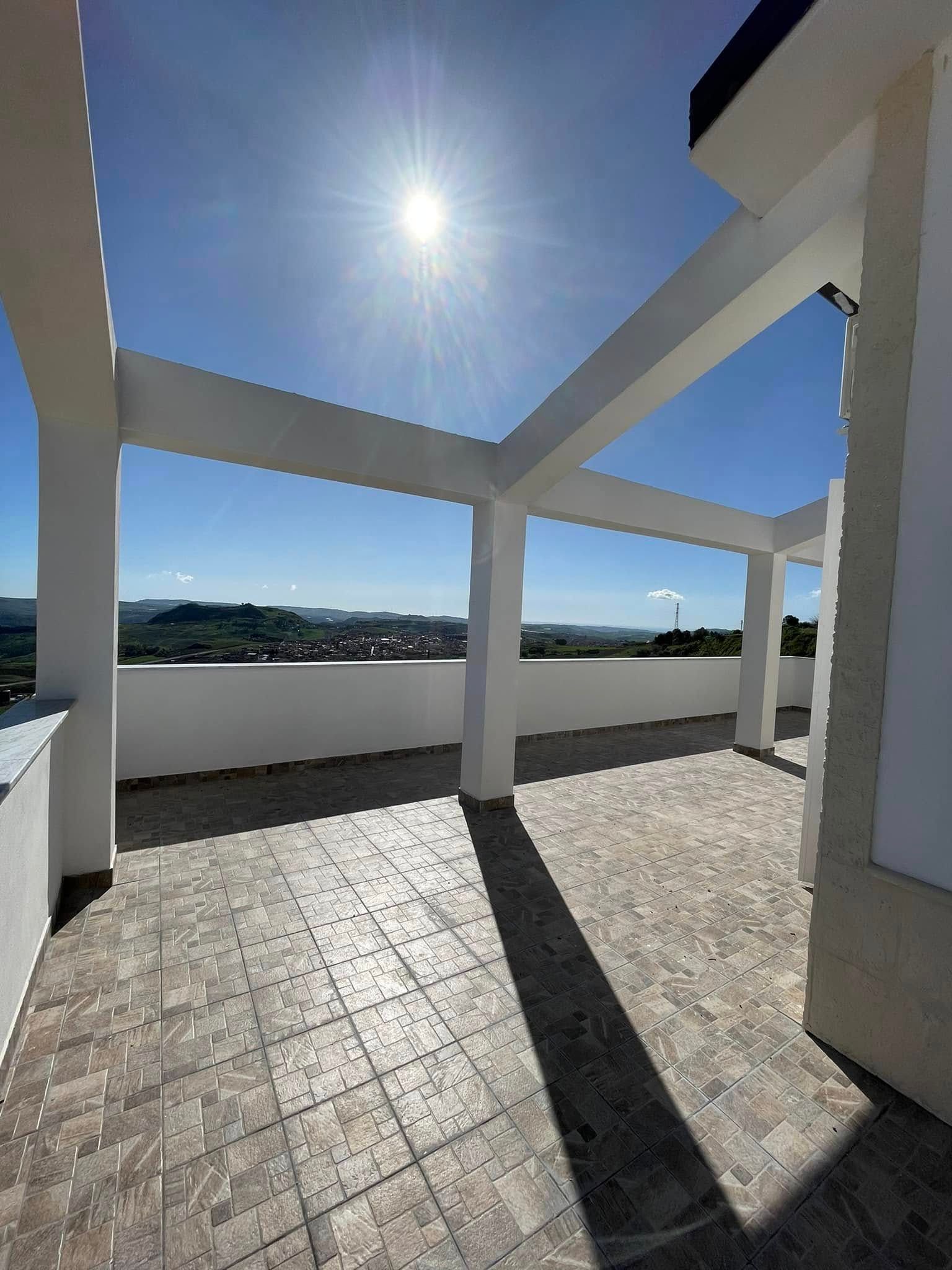 Terrazza panoramica soleggiata con pavimento piastrellato, pilastri bianchi e vista sull'oceano in lontananza.